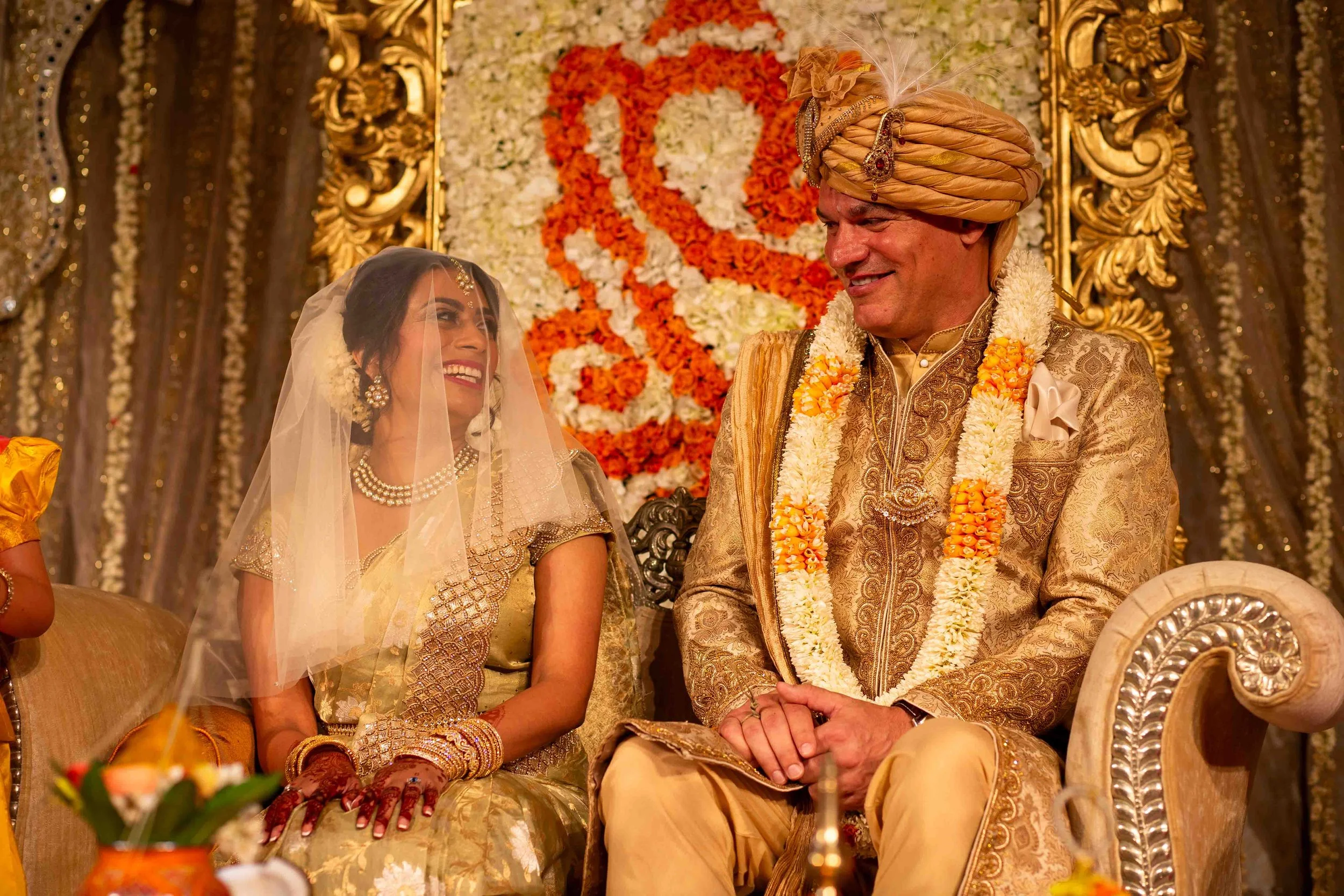 A bride and groom sit on a decorated stage during a traditional Indian wedding. The bride is wearing a gold saree, jewelry, and a veil, while the groom is dressed in an ornate golden sherwani with a turban. They are smiling at each other as they hold