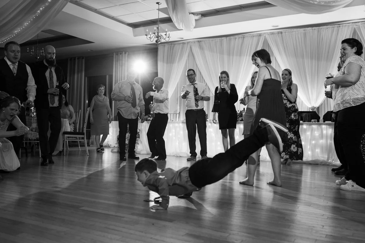 A young boy in a dress shirt and dark pants is doing a push-up on the dance floor at a celebration or party. People in formal attire are standing and sitting around, smiling and watching, in a decorated banquet hall.