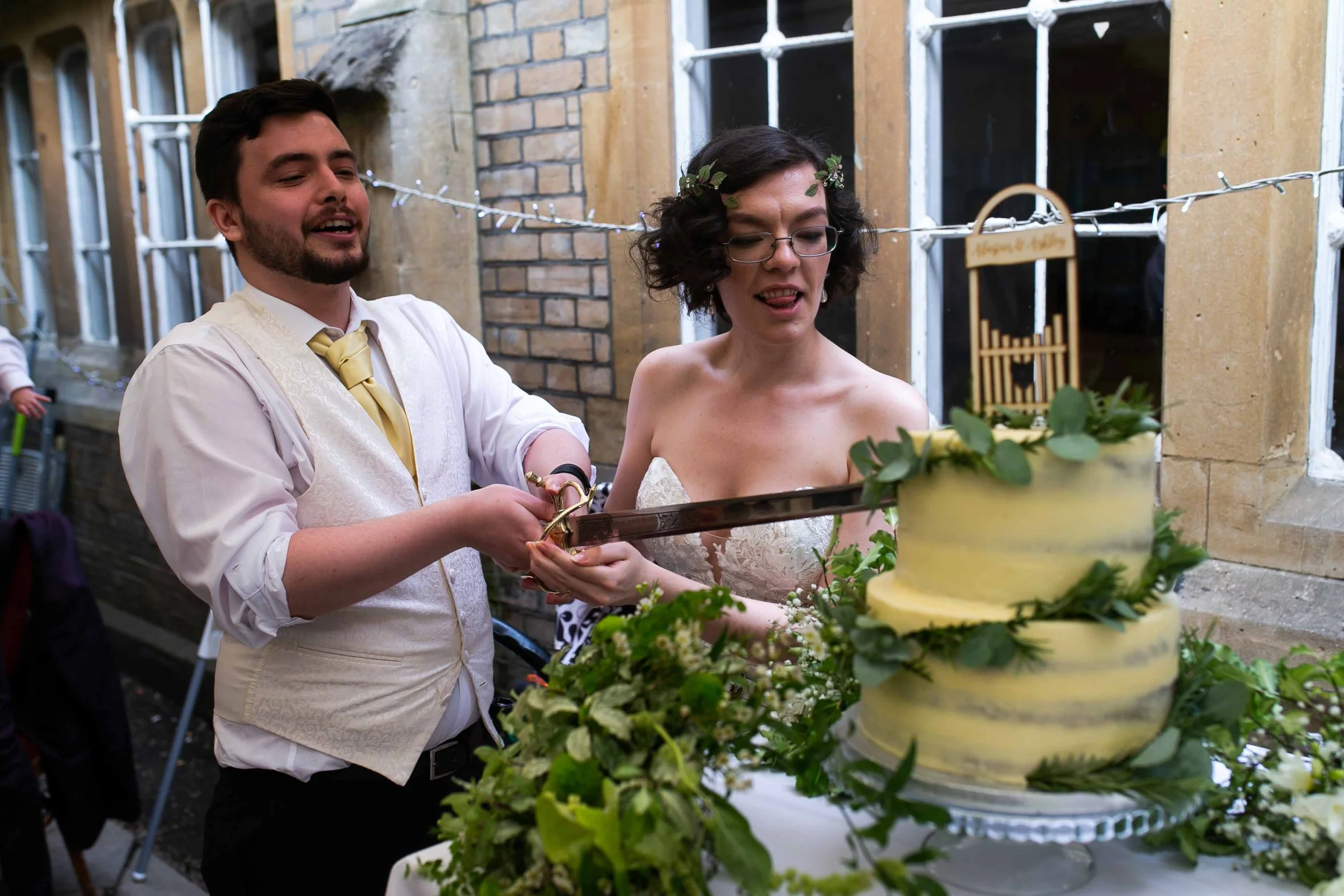 A man and woman are cutting a wedding cake together, surrounded by greenery and decorations, during an outdoor celebration at night.