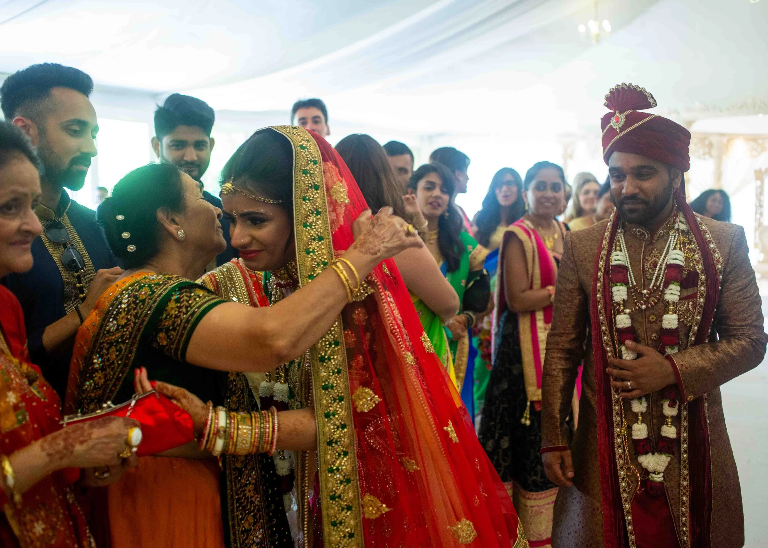 Indian bride in a red and gold traditional wedding outfit being greeted by an elderly woman, surrounded by guests in colorful attire at a wedding celebration.