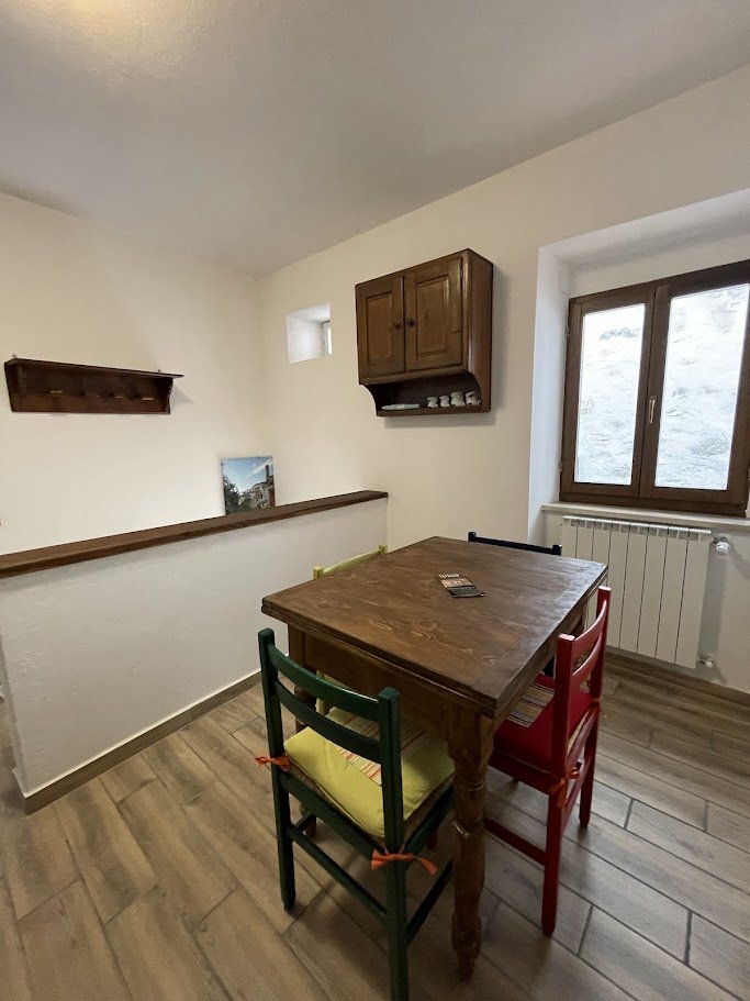Interno di una stanza con un tavolo in legno e quattro sedie colorate. C'è una finestra alla destra e un termosifone sotto di essa. Un mobile sospeso con tazze e un piccolo scaffale decorativo sono appesi al muro. Il pavimento è in legno chiaro.
