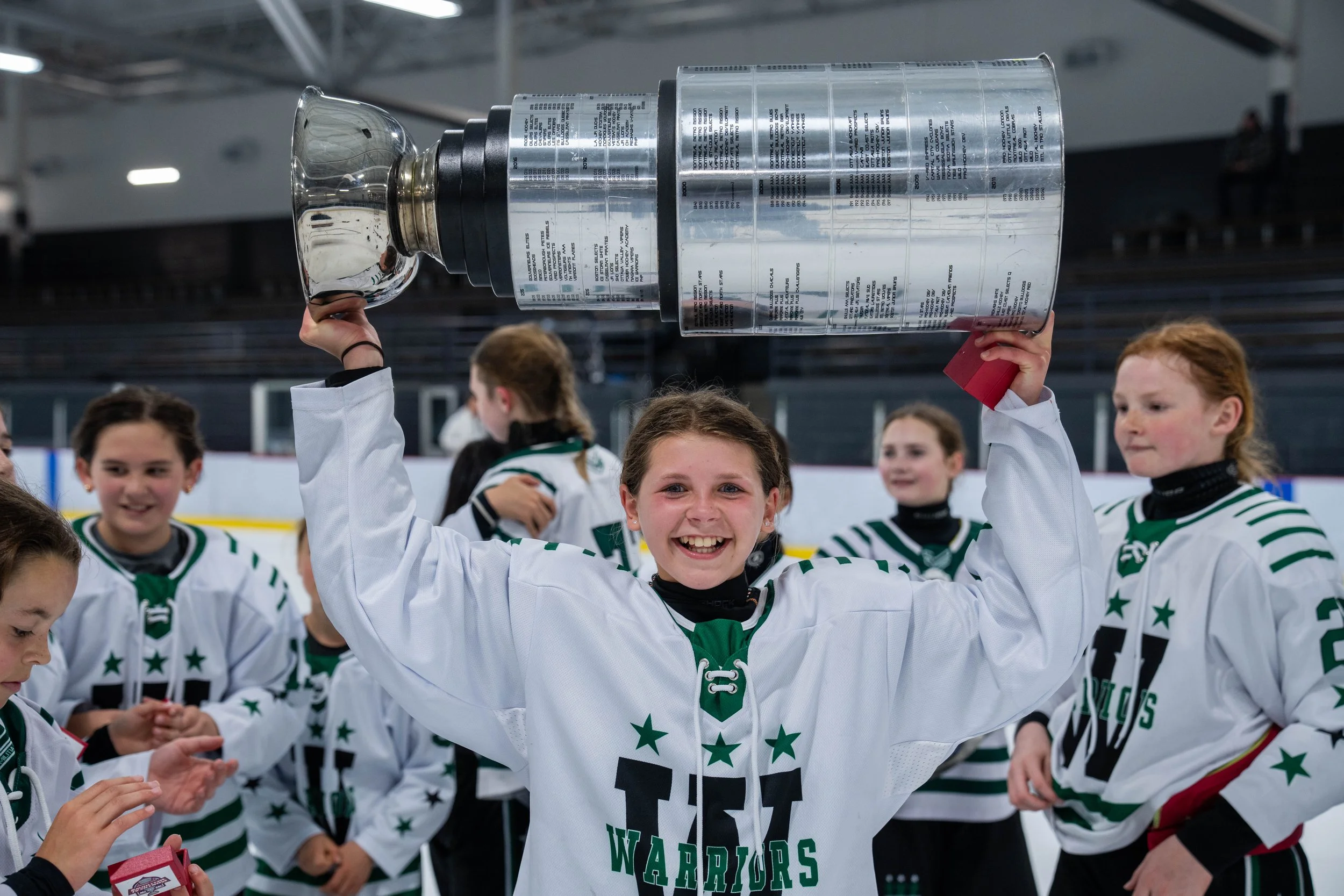 Youth female hockey player holding a trophy over her head, celebrating with teammates on an ice rink.