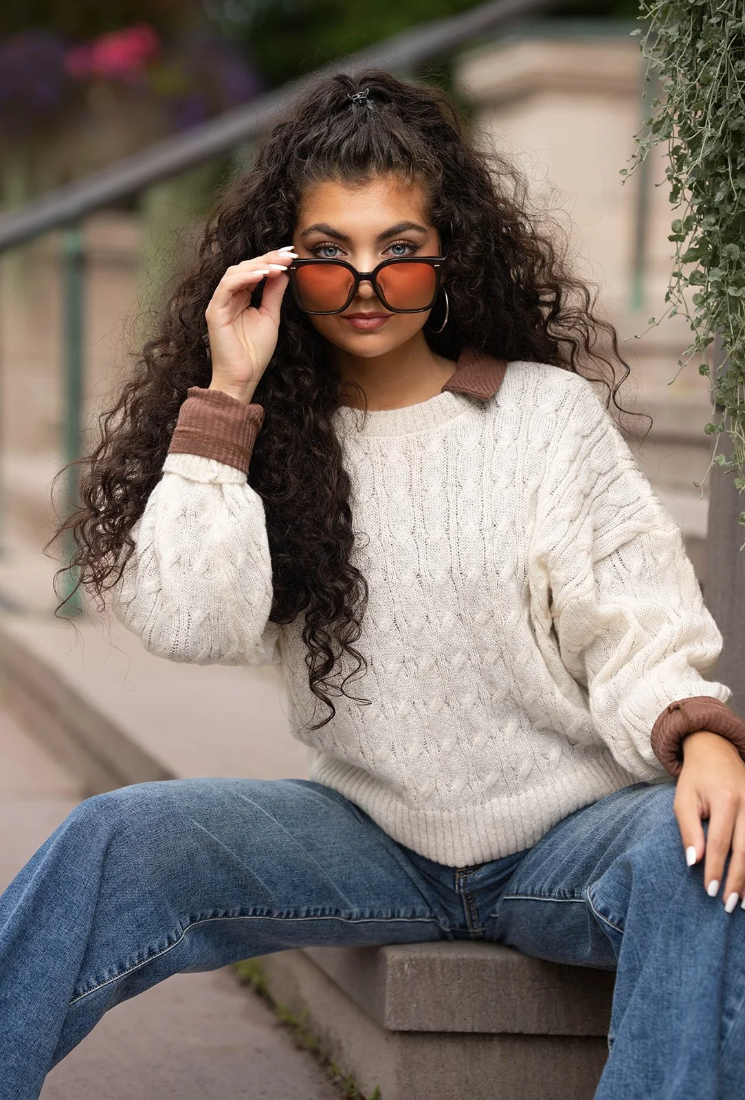 Woman with curly hair wearing sunglasses, a cream cable-knit sweater, and jeans, sitting on outdoor steps.