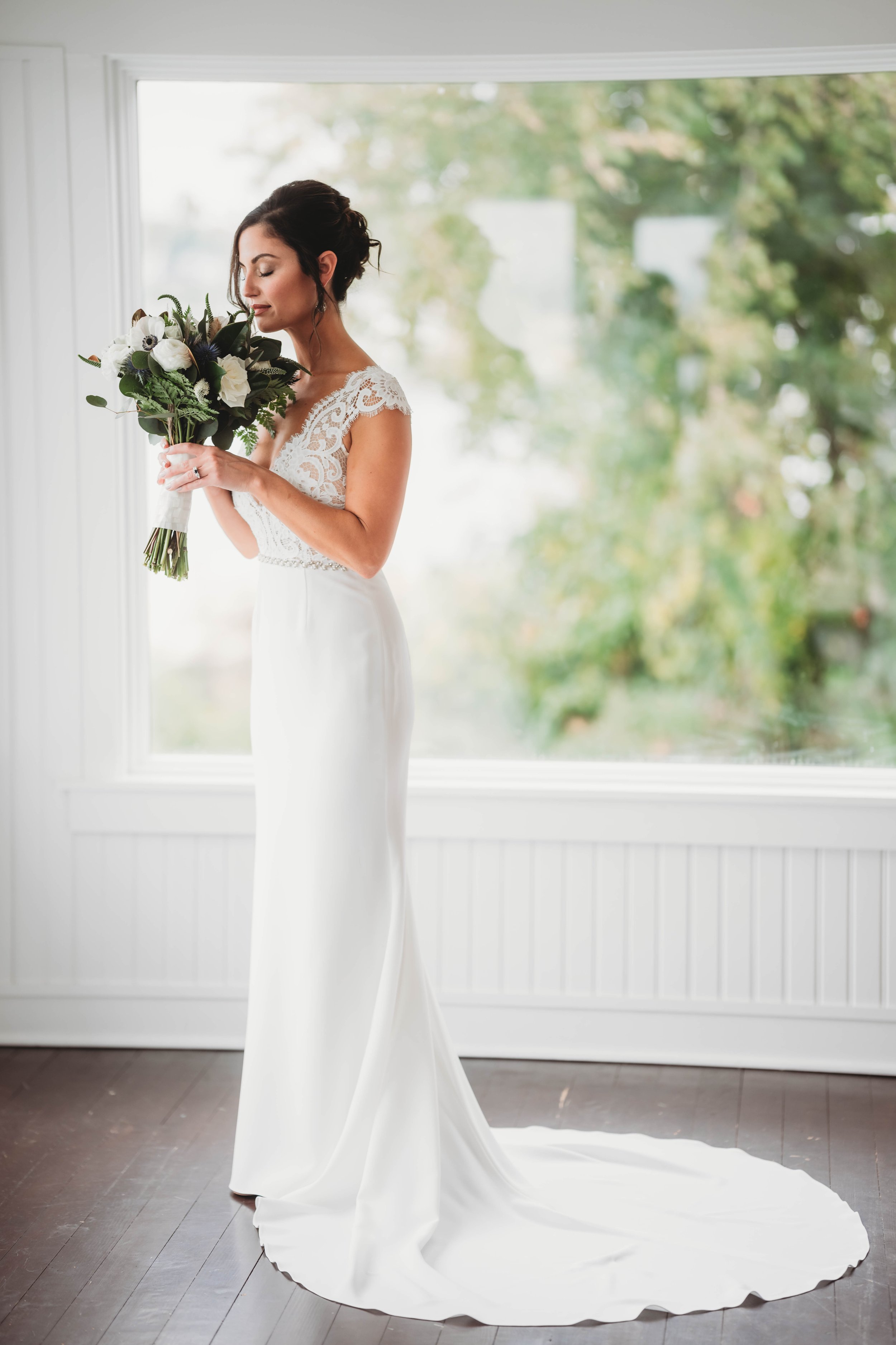 Bride holding bouquet standing indoors by window with greenery outside.
