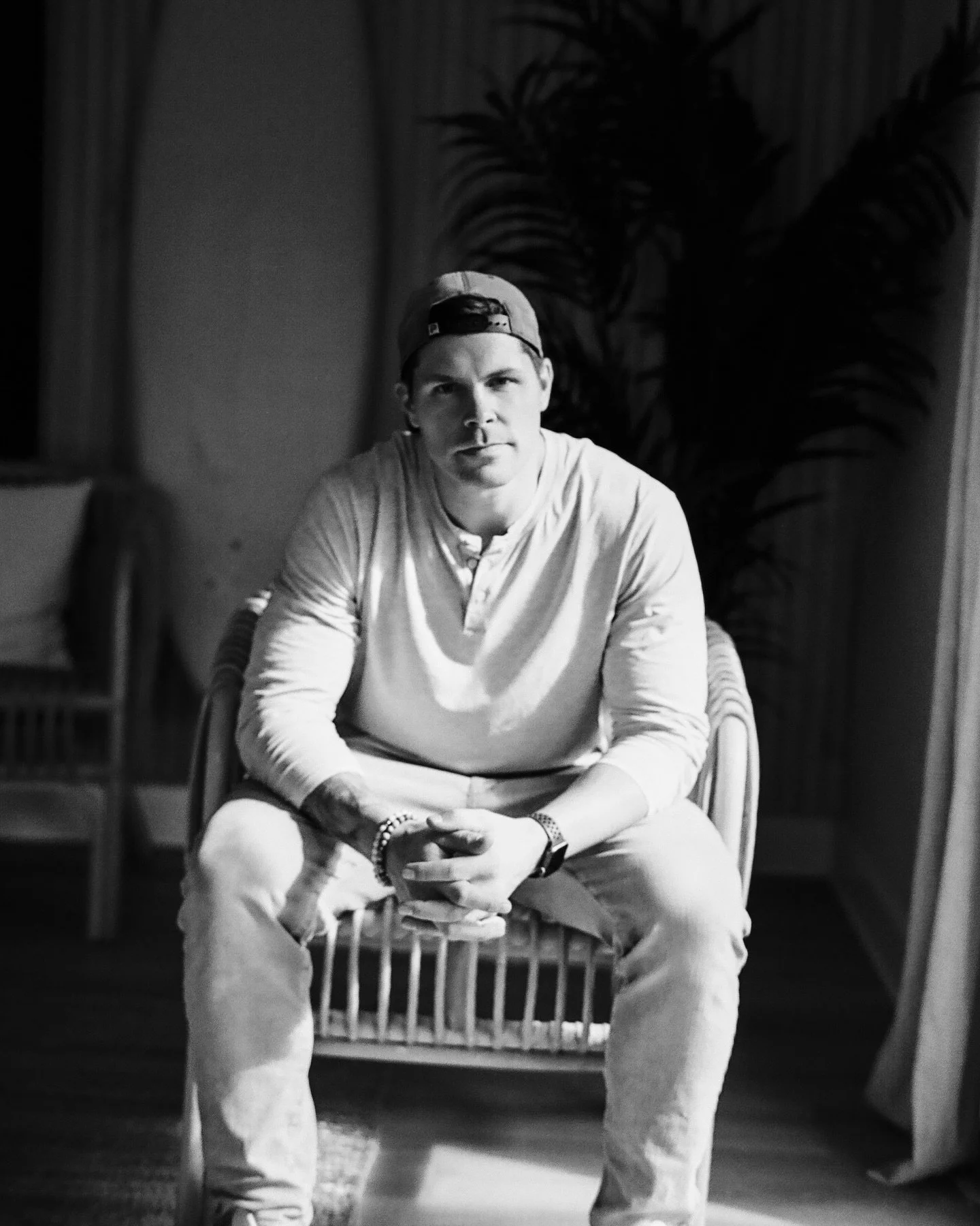 Black and white photo of a man sitting on a chair indoors, wearing a cap and casual clothing, with a surfboard and plant in the background.