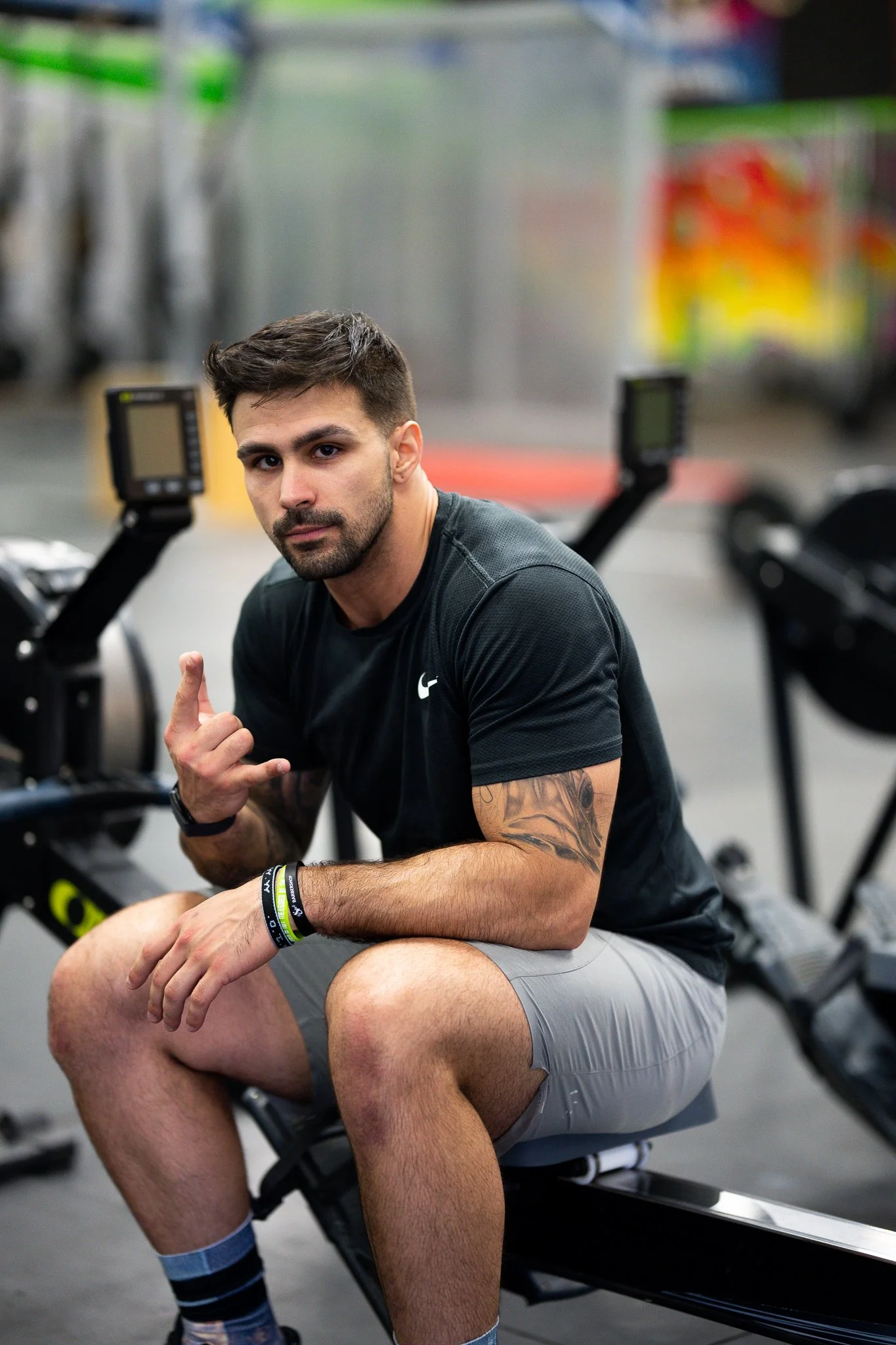 Man sitting on rowing machine in gym, showing hand gesture, wearing black shirt and gray shorts, with tattooed arm.