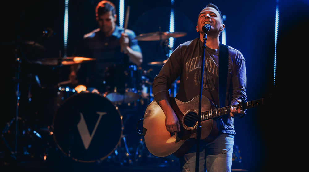 A singer playing an acoustic guitar and singing into a microphone on stage, with a drummer performing in the background, illuminated by stage lights.