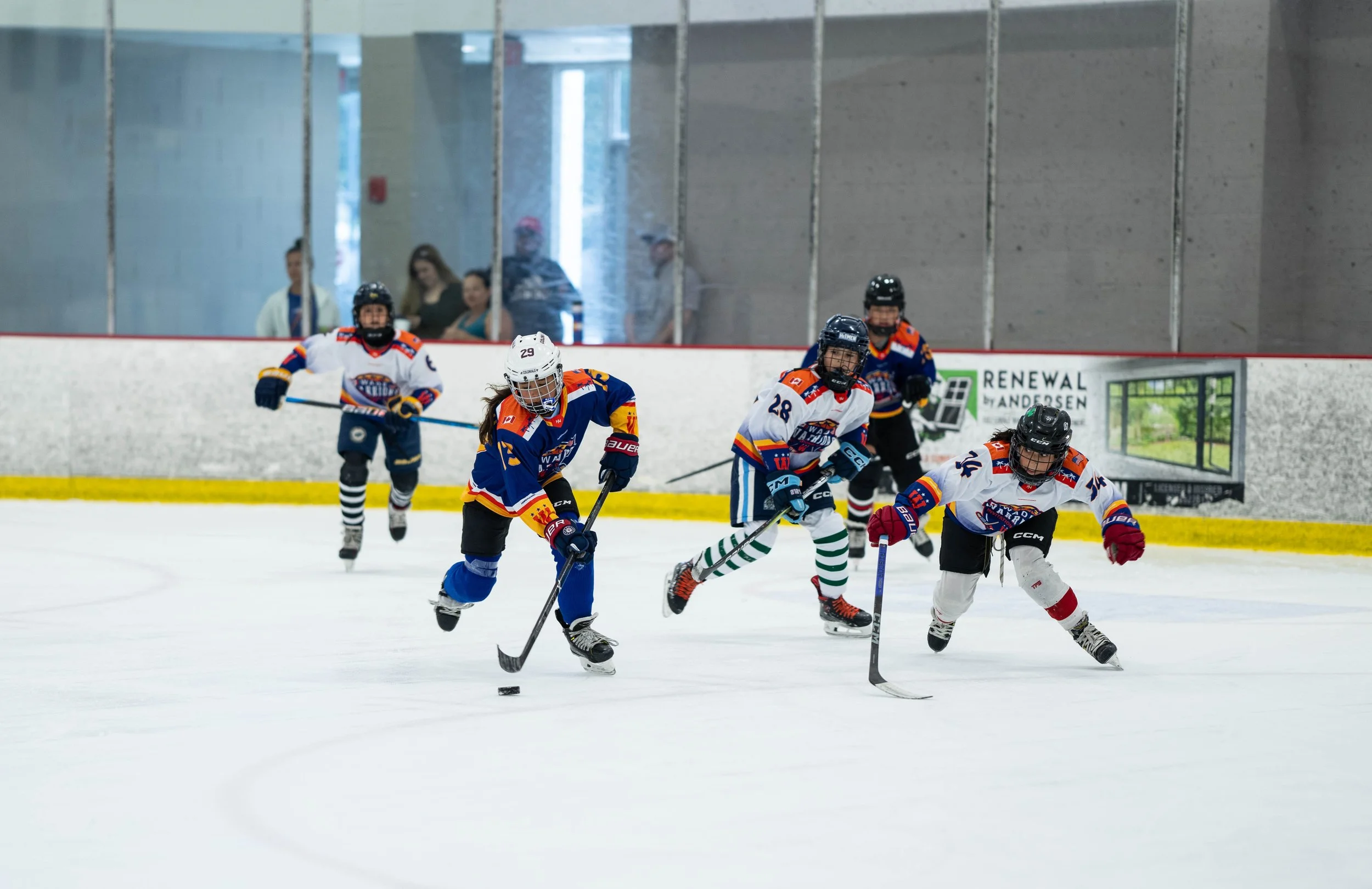Youth hockey players in action on an ice rink, wearing colorful uniforms and helmets, competing in a game while spectators watch from behind the glass.