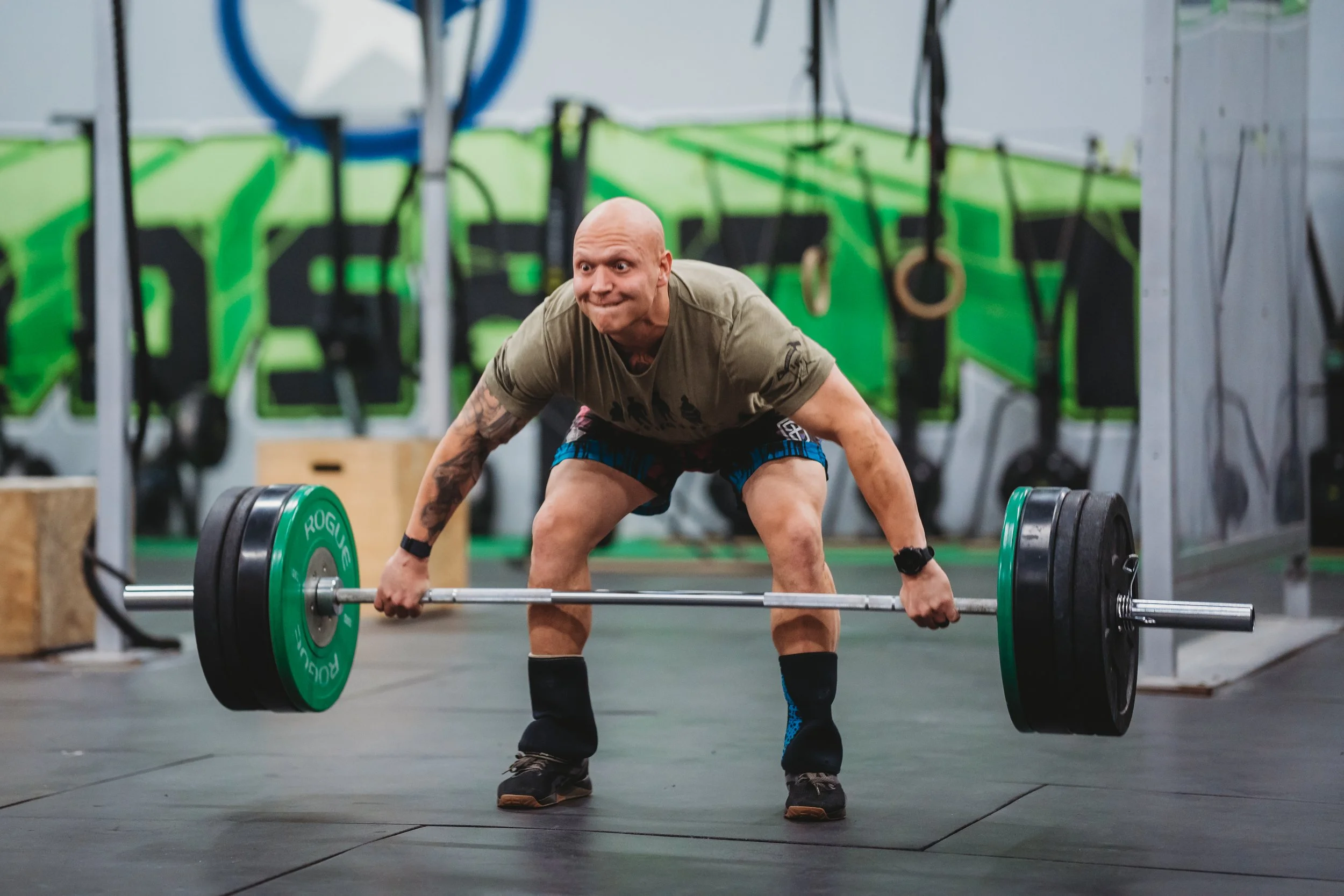 Man performing a deadlift in a gym with green wall, wearing a tan shirt and black gear, using a barbell with green and black weights.