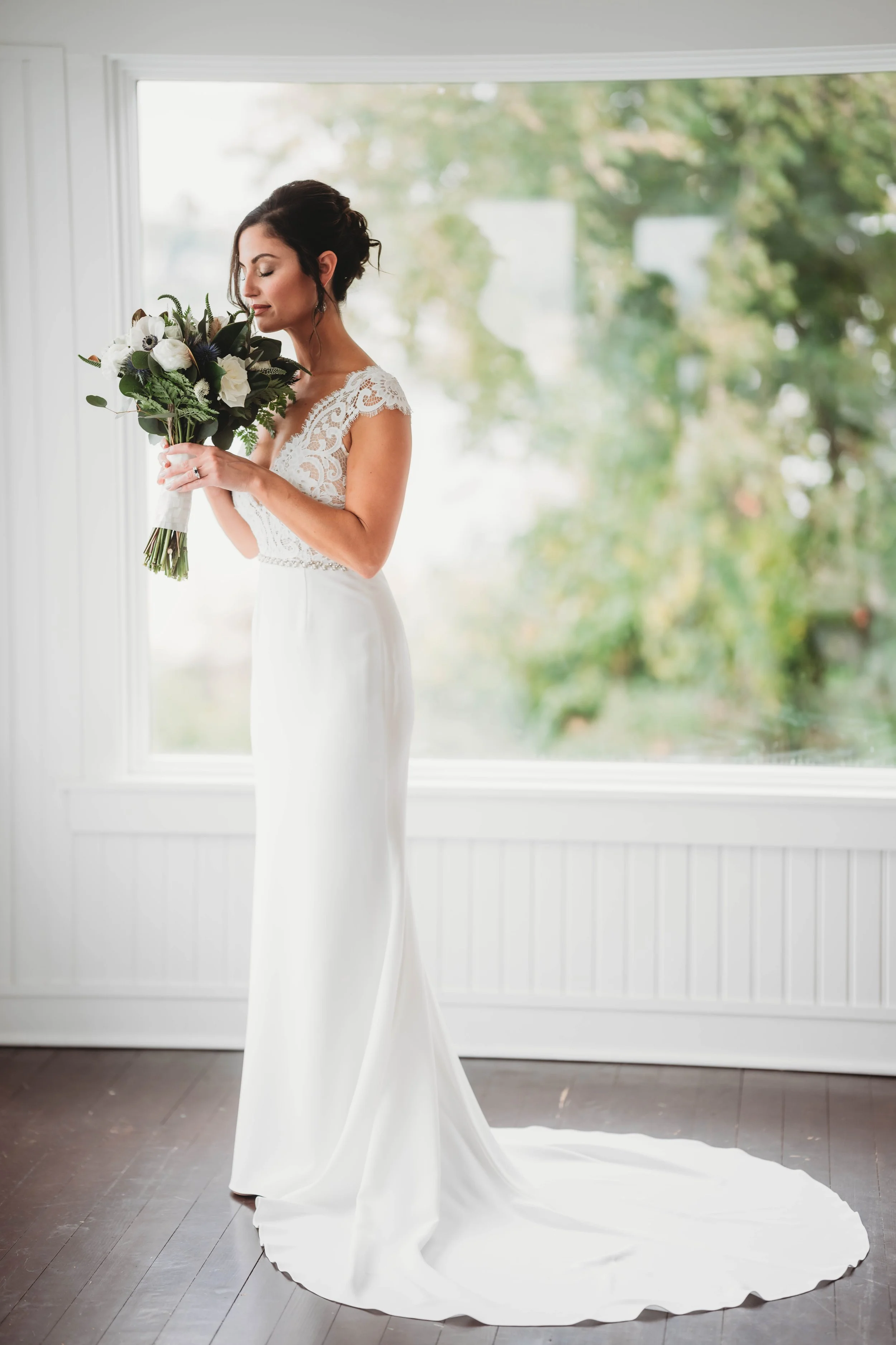 Bride in an elegant white gown holding a bouquet, standing by a large window with green foliage in the background.