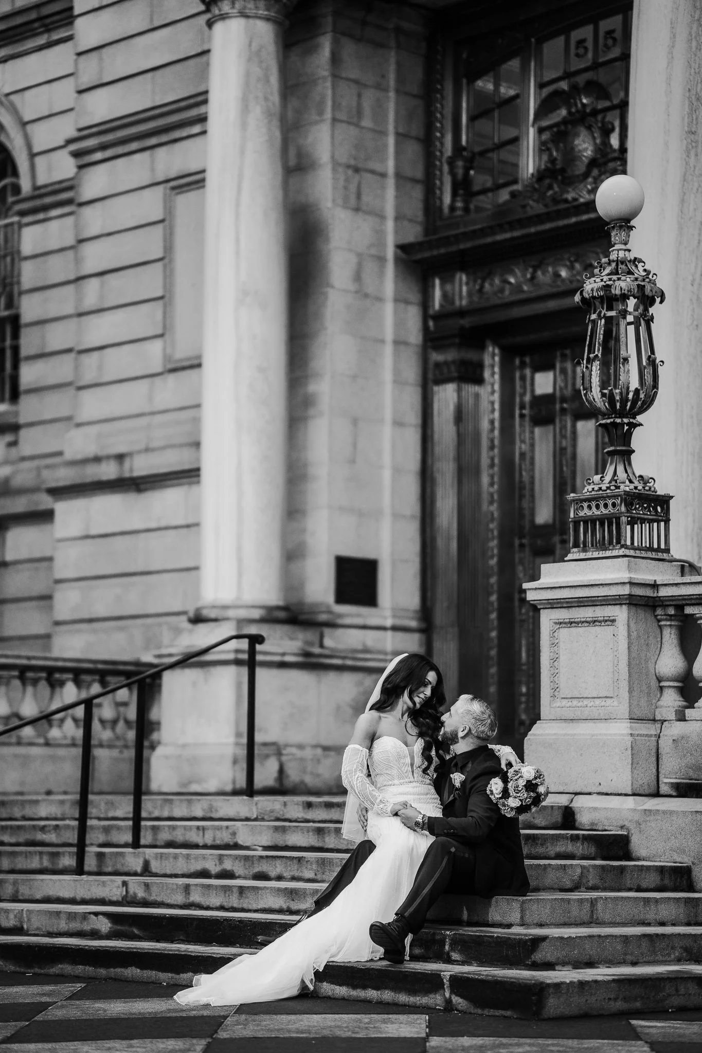 Bride and groom sitting on steps, smiling at each other, in front of a large ornate building. The bride is wearing a wedding dress, and the groom is in a suit, holding a bouquet.