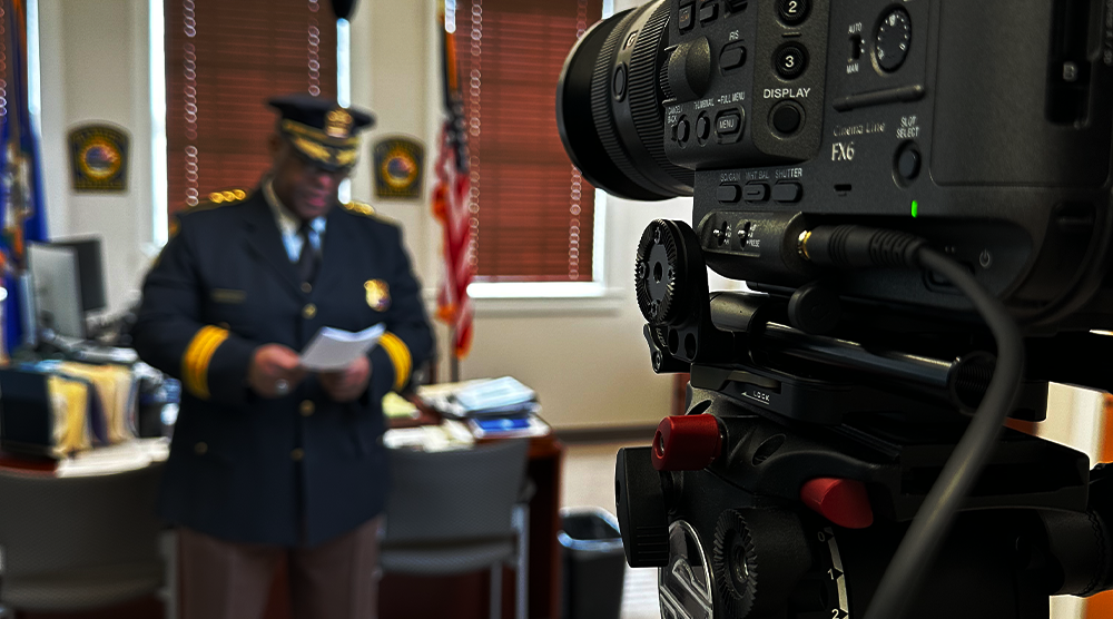 Close-up of a professional video camera focusing on a uniformed officer in an office with flags and badges in the background.