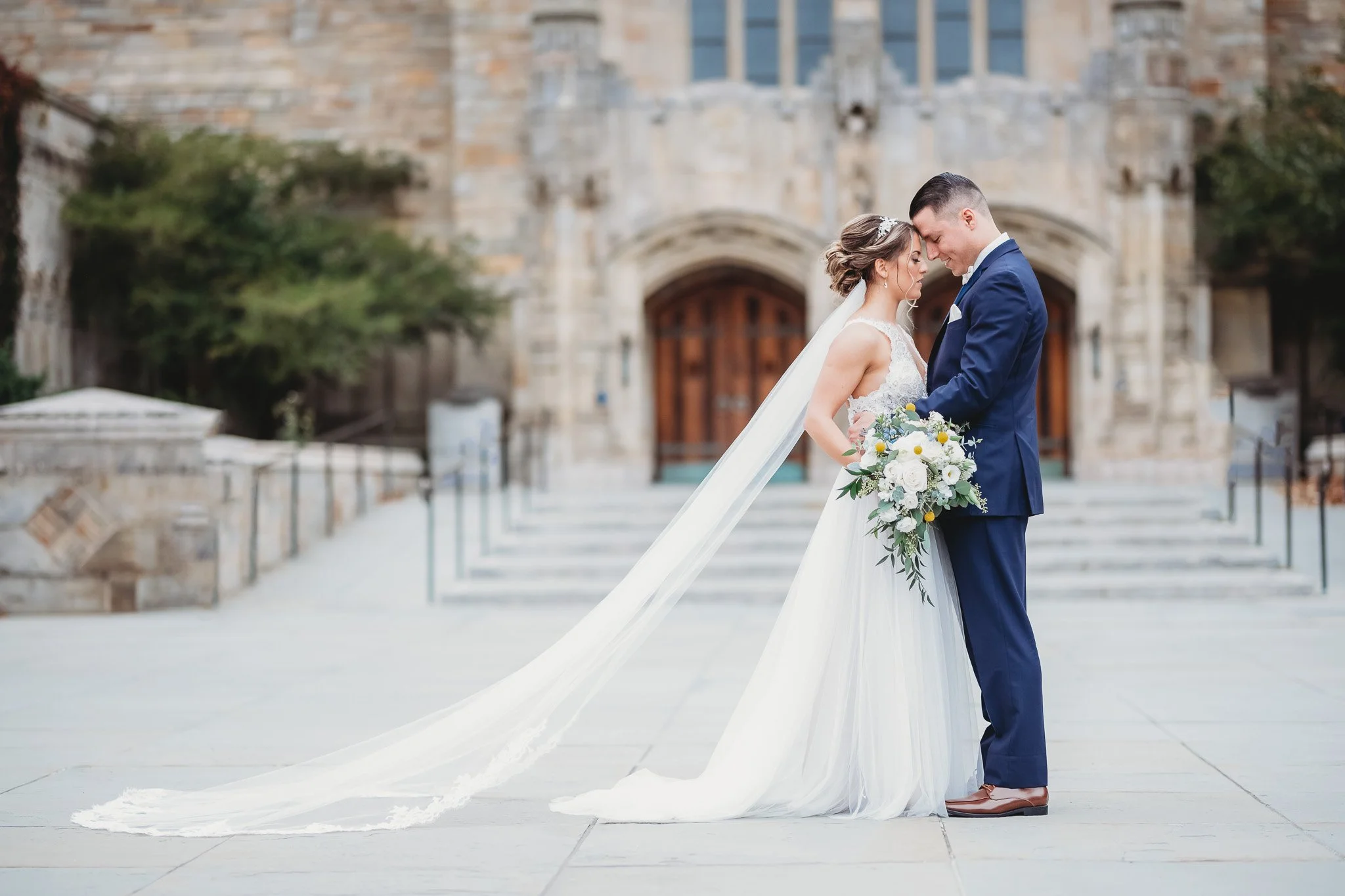 Bride and groom standing close, embracing, with bride's long veil flowing behind, holding a bouquet, in front of a historic building.