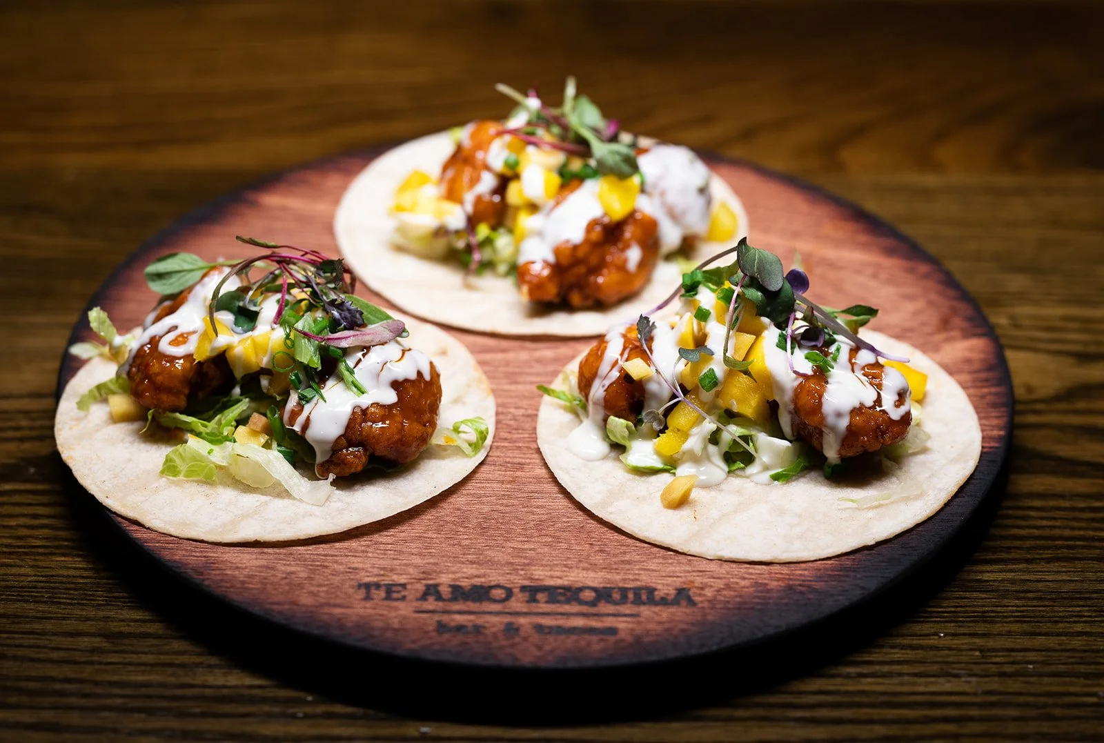 Three tacos on a wooden platter, topped with fried chicken, drizzle of creamy sauce, diced mango, microgreens, and chopped lettuce.