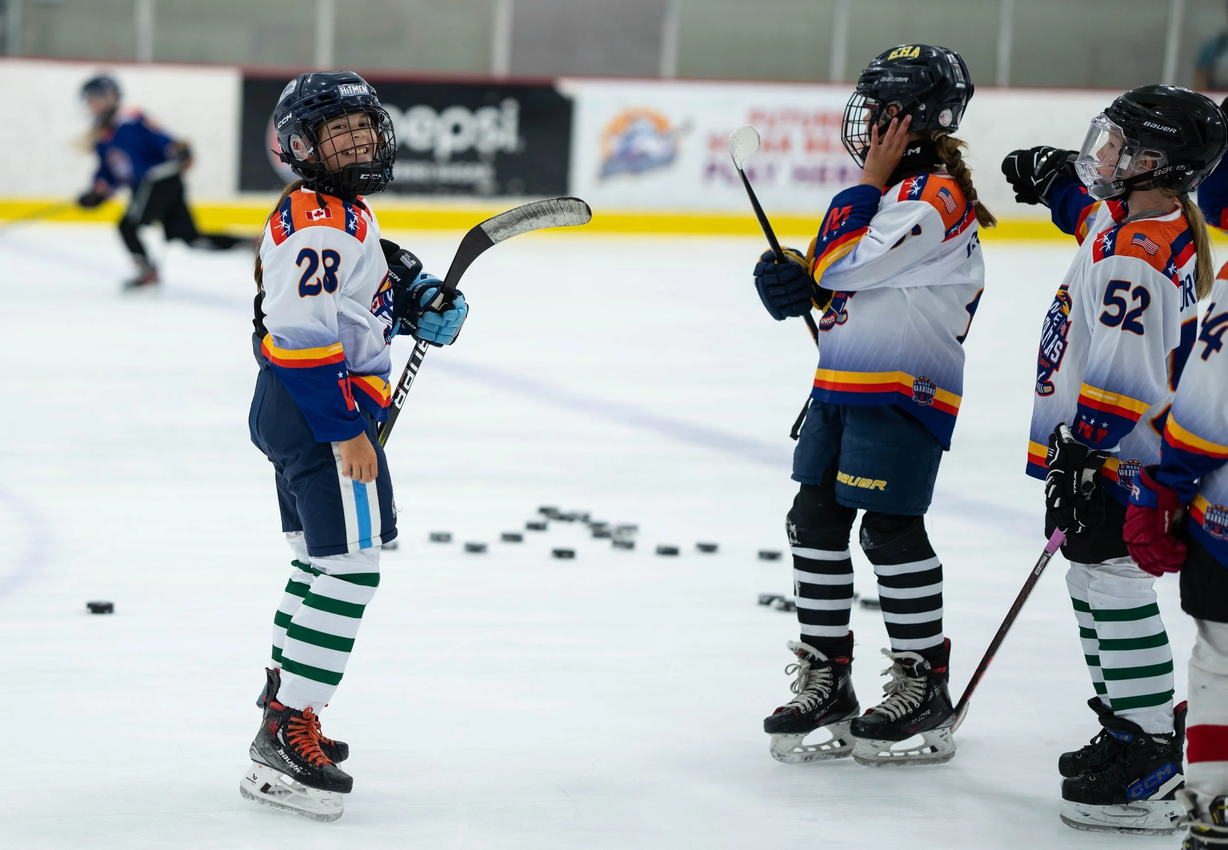 Young hockey players on ice rink in uniforms and helmets holding hockey sticks, with pucks scattered on the ice.