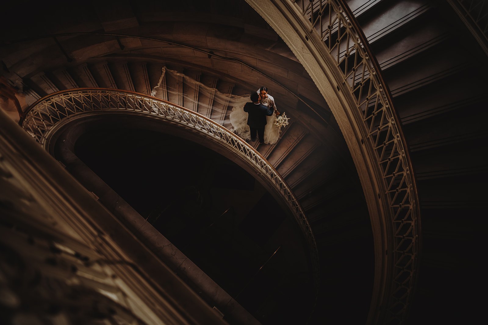Aerial view of a couple in wedding attire on a spiral staircase, with intricate railings and dramatic lighting.