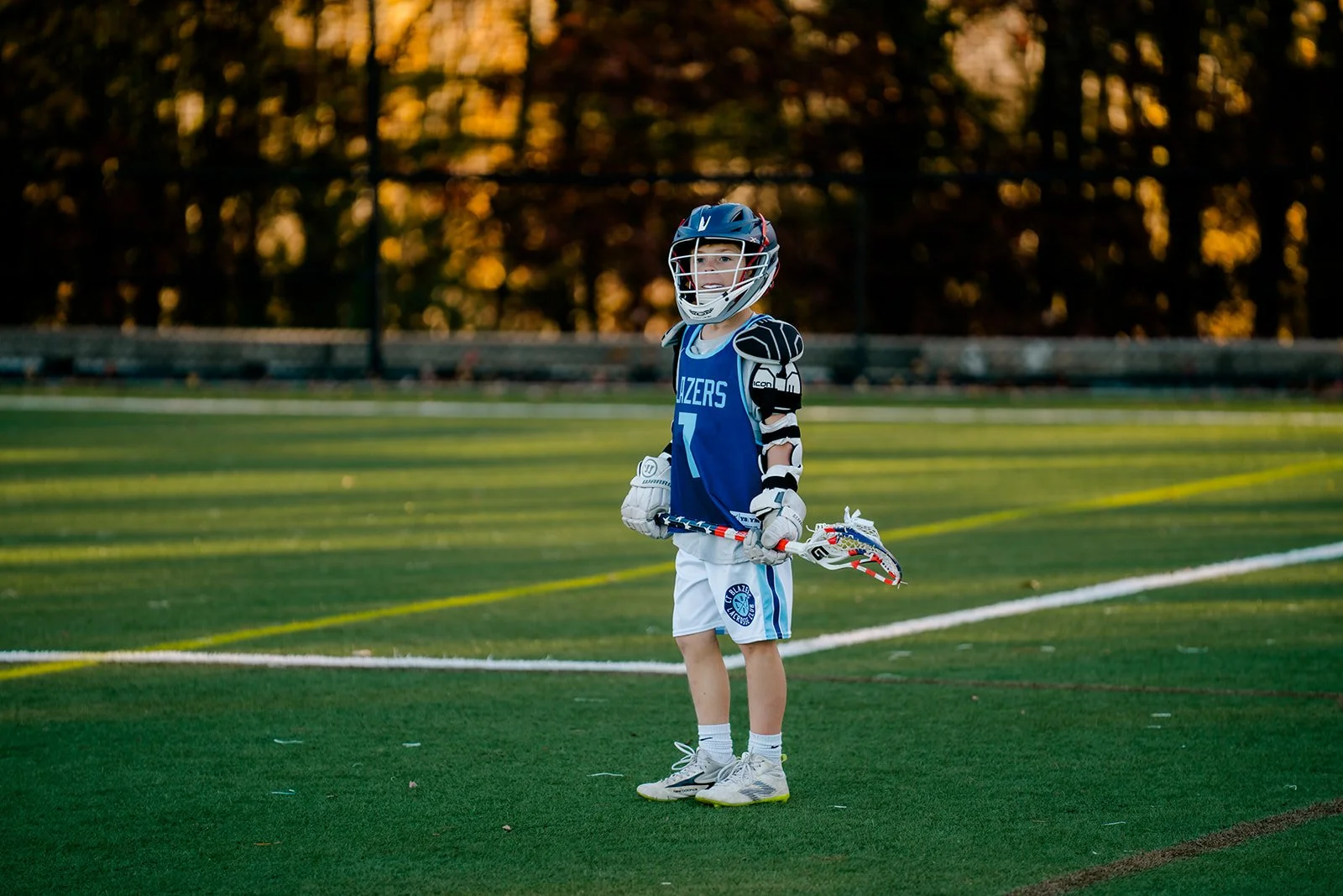 A young lacrosse player in a blue jersey and helmet standing on a sports field with a lacrosse stick.