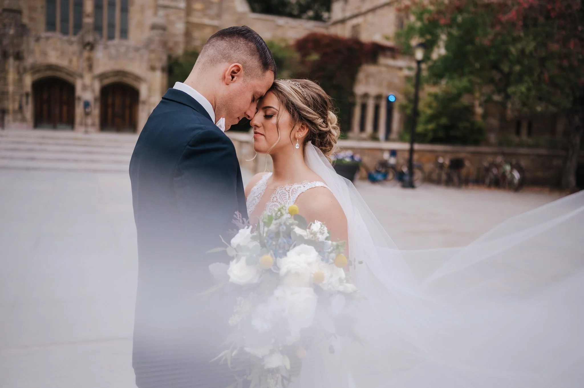 Bride and groom embracing outdoors, with a bouquet of flowers and a flowing veil, in front of a historic stone building.