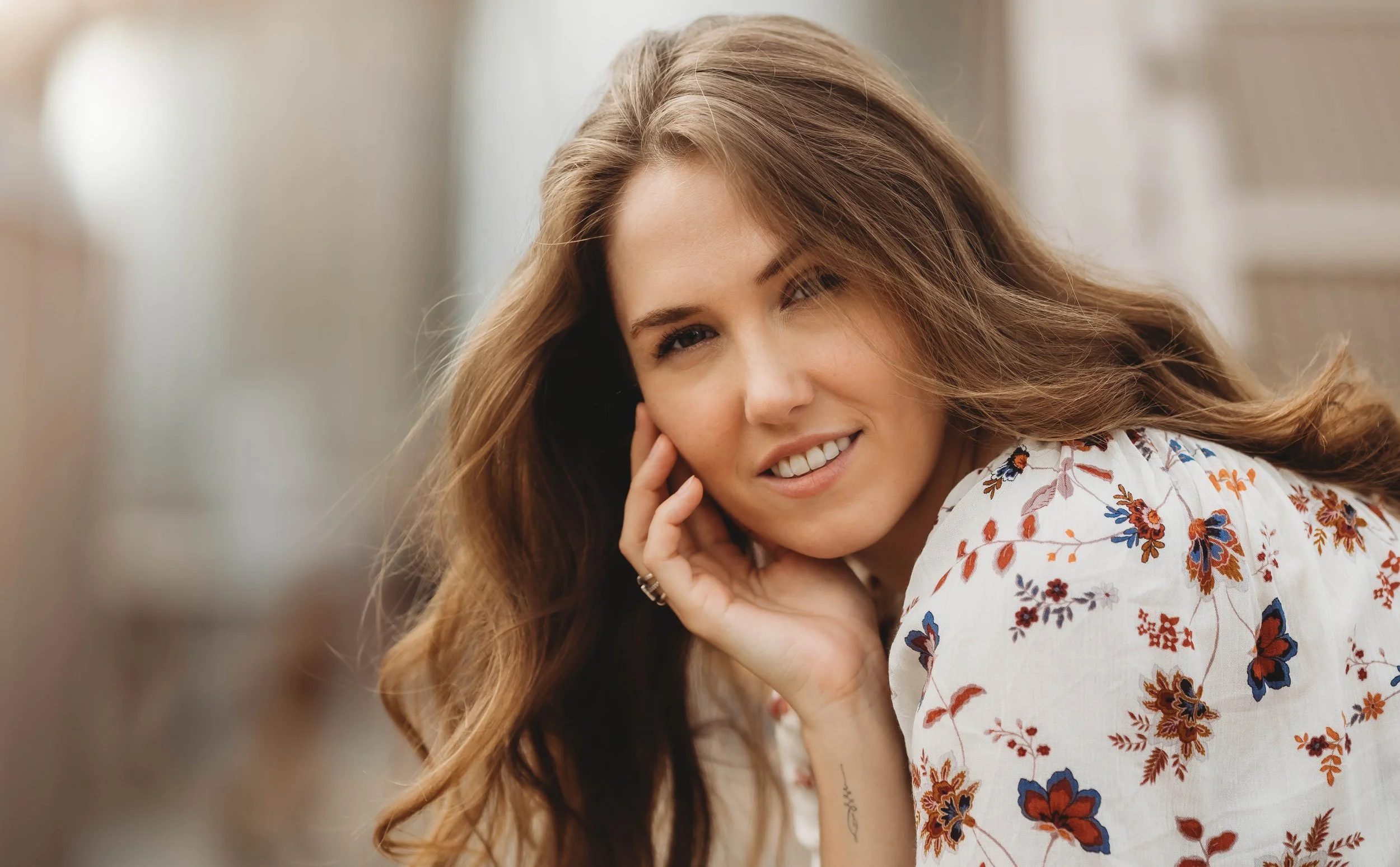 A woman with long brown hair smiles gently, resting her face on her hand. She wears a white top with colorful floral patterns.