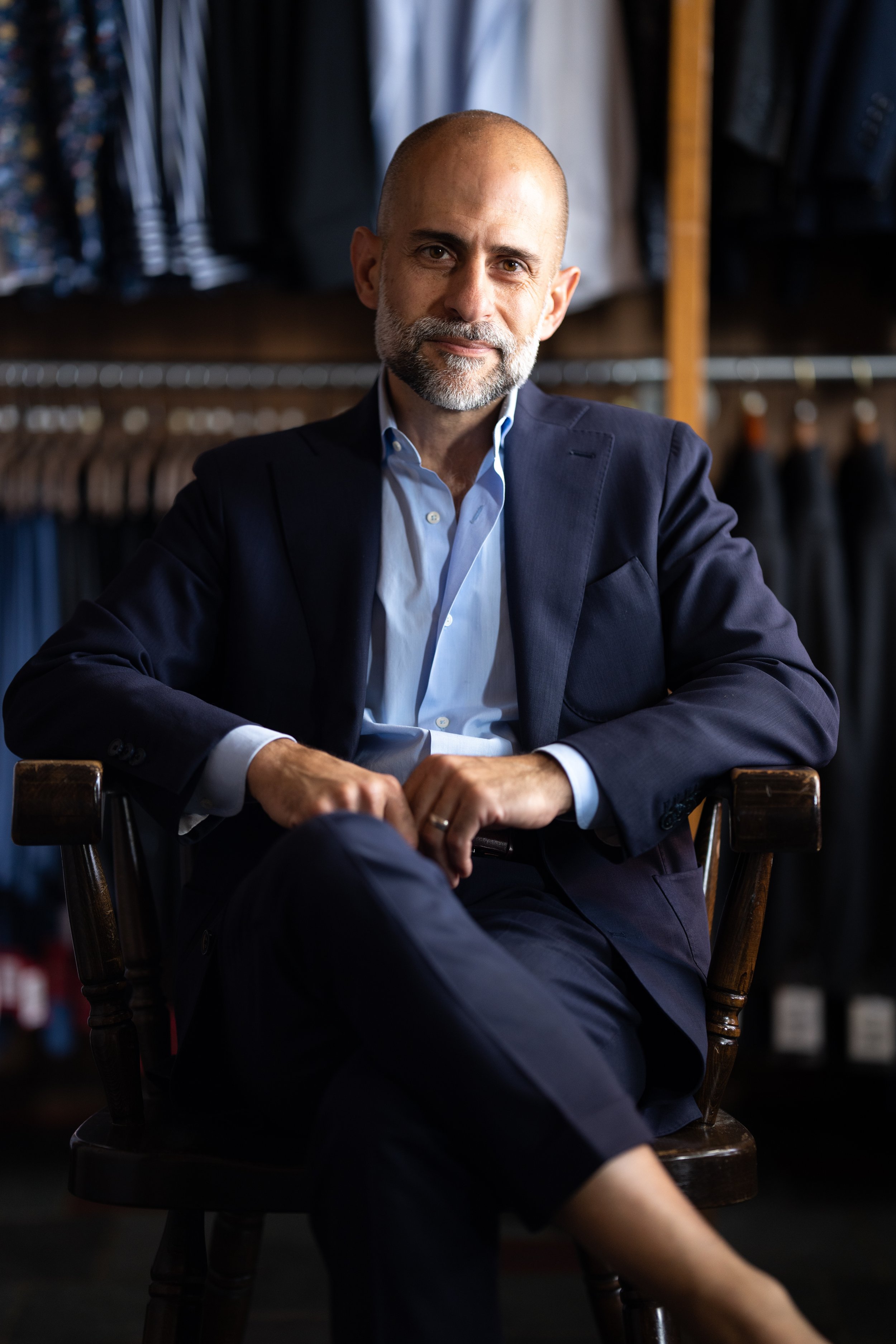 Man in a suit sitting in a chair with clothes on hangers in the background.