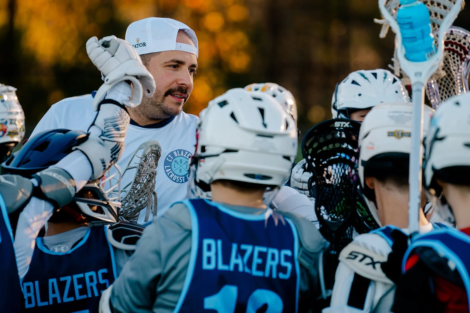 Lacrosse coach talking to a team of young players wearing helmets and jerseys labeled "Blazers," holding lacrosse sticks.