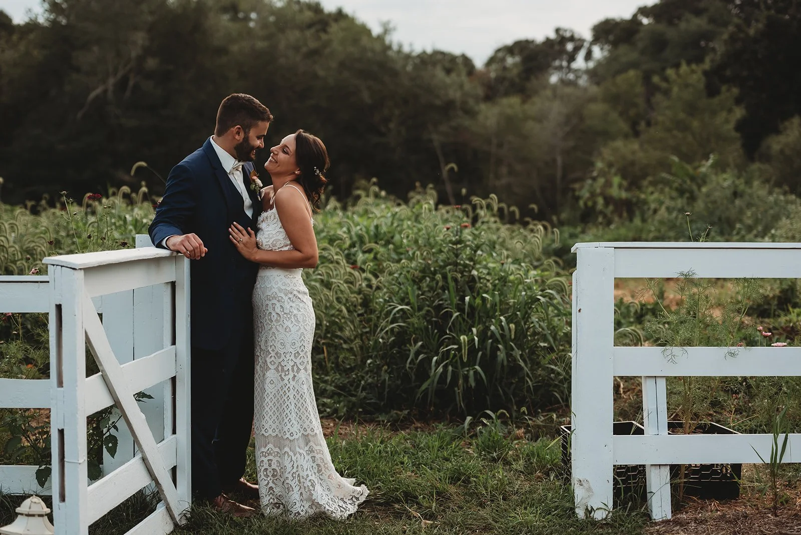 A couple in formal attire stands by a white wooden gate in an outdoor setting, surrounded by greenery. The woman wears a white lace dress, and the man wears a dark suit. They appear to be smiling and embracing each other.