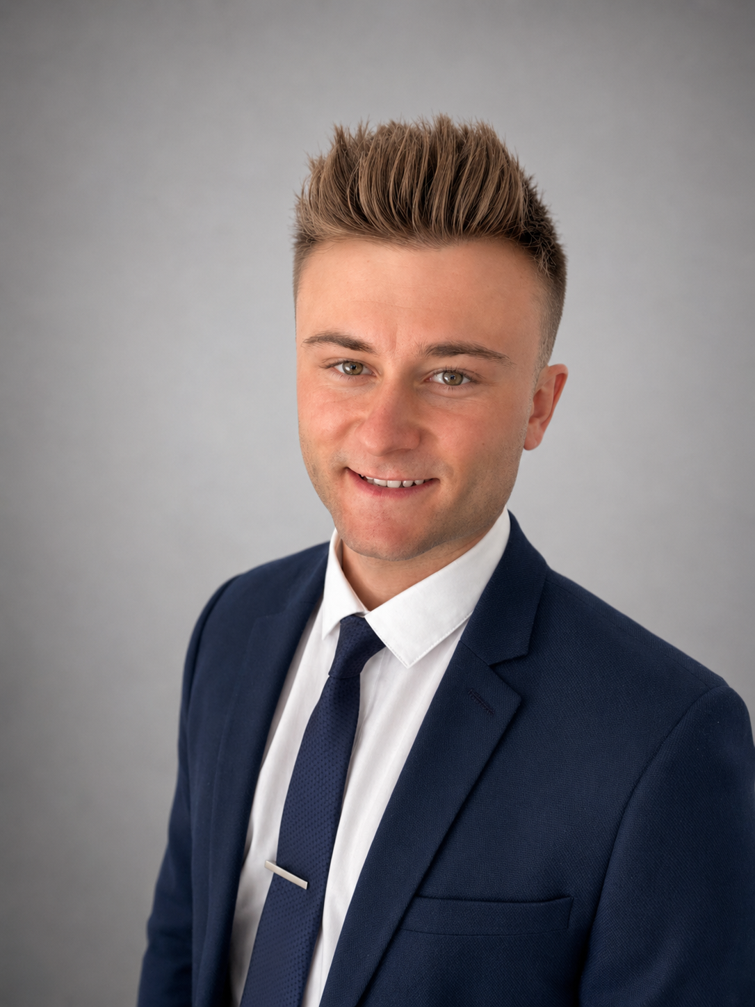 A young man in a navy blue suit, white shirt, and navy blue tie smiling at the camera with a light gray background.