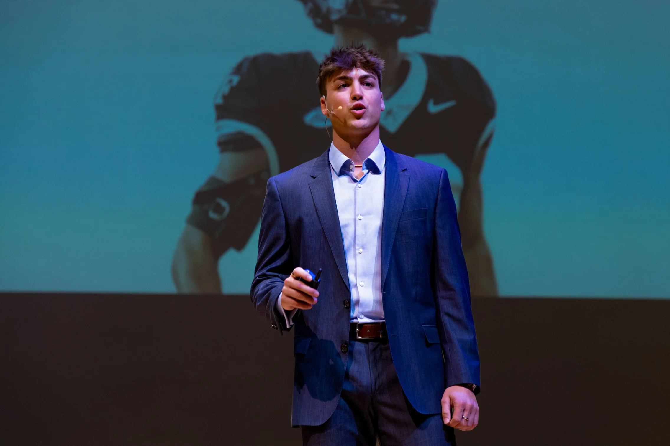 Young man in a blue suit and white shirt smiling outdoors with a blurred building and trees in the background.