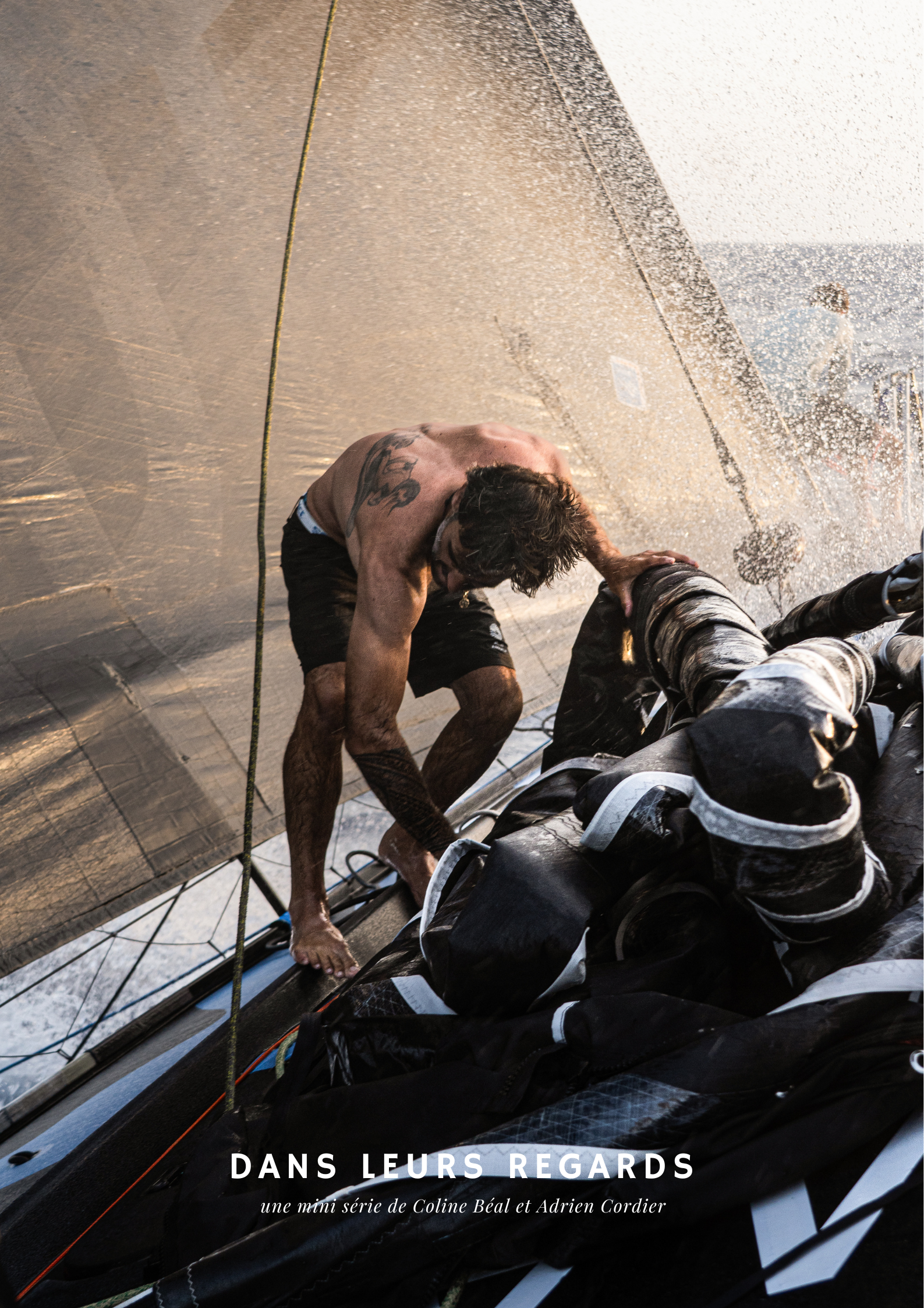 Un marin nu, en short, se relève sur un bateau de compétition après une mer agitée, avec des voiles en arrière-plan, portant un tatouage sur le bras gauche.