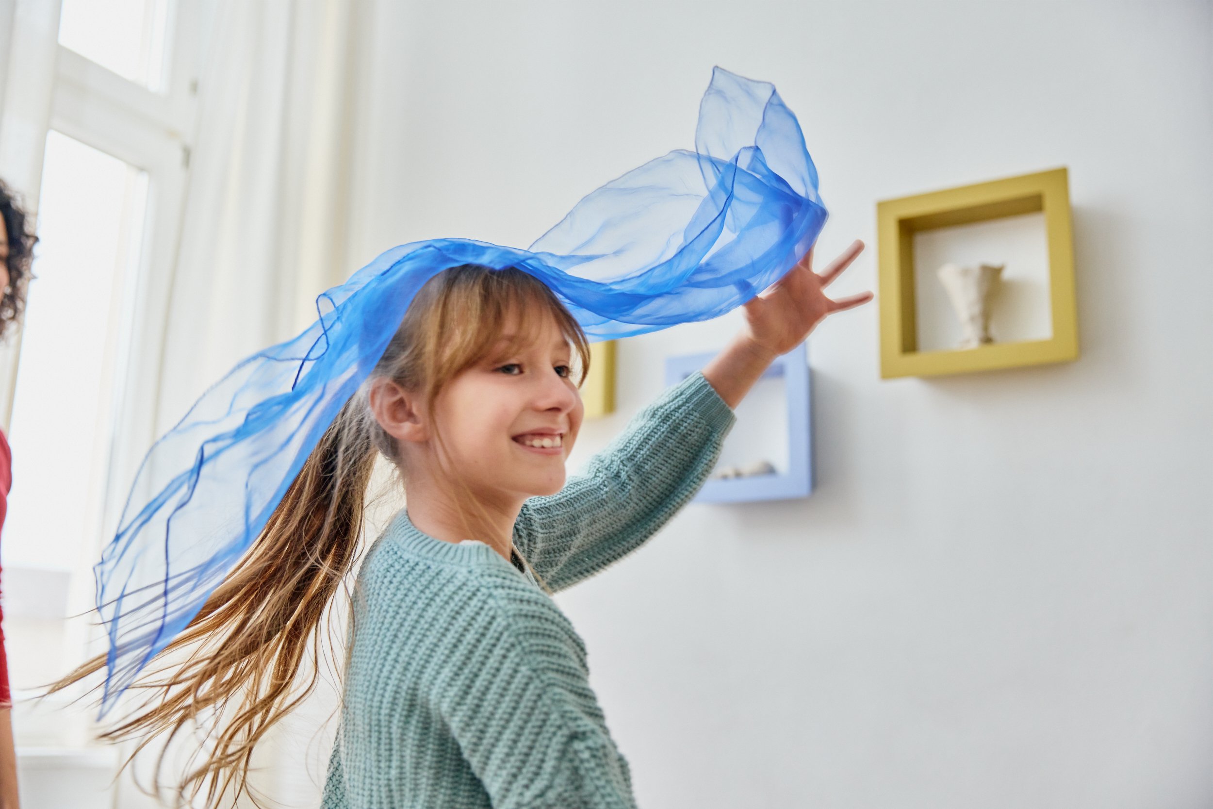 Ein lachendes Mädchen mit langem Haar trägt ein blaues Tuch auf dem Kopf in einem Raum mit weißen Wänden und bunten Bilderrahmen.