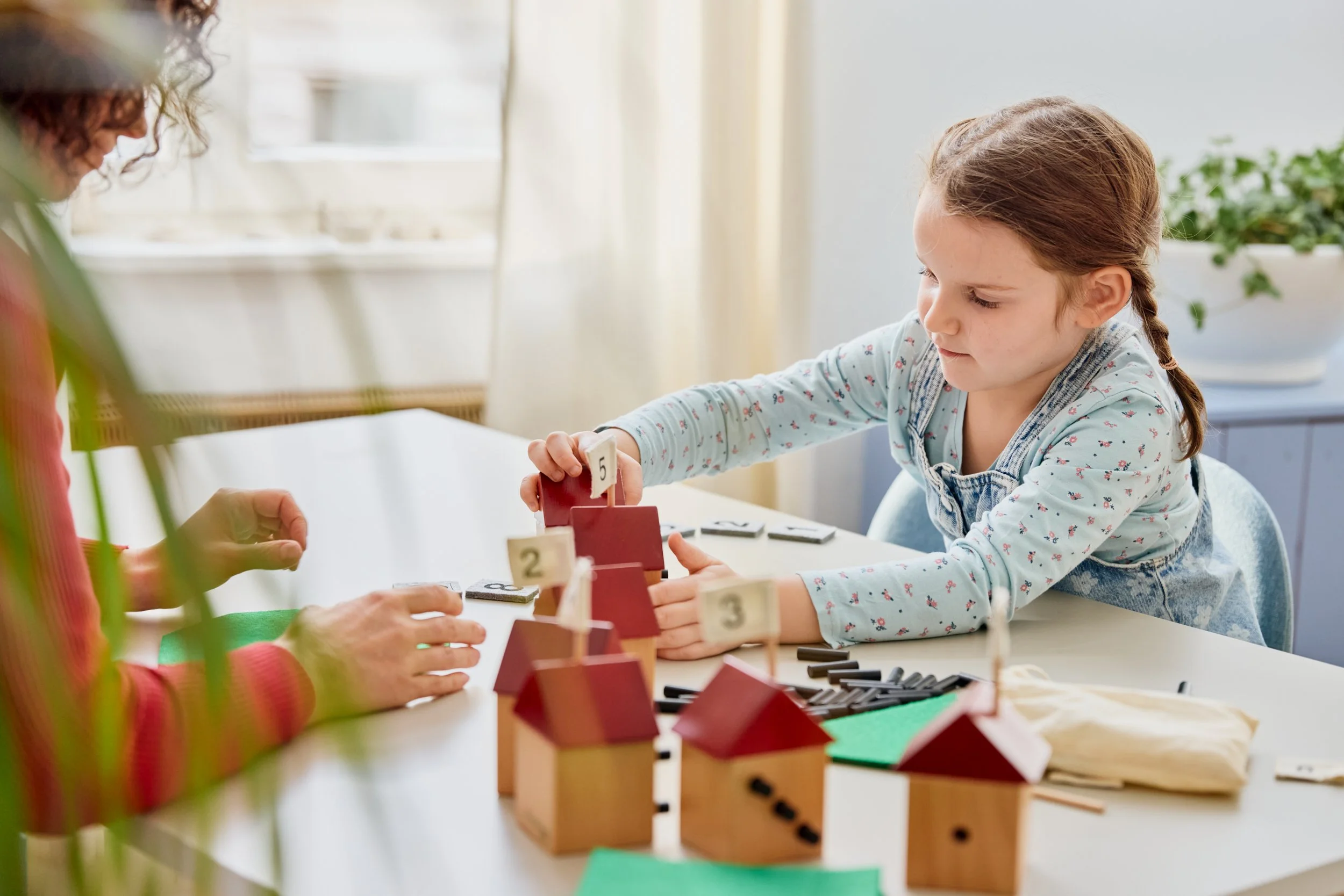 Ein Mädchen spielt mit Holzhaus-Spielen und Zahlen, während ein Erwachsener neben ihr sitzt.