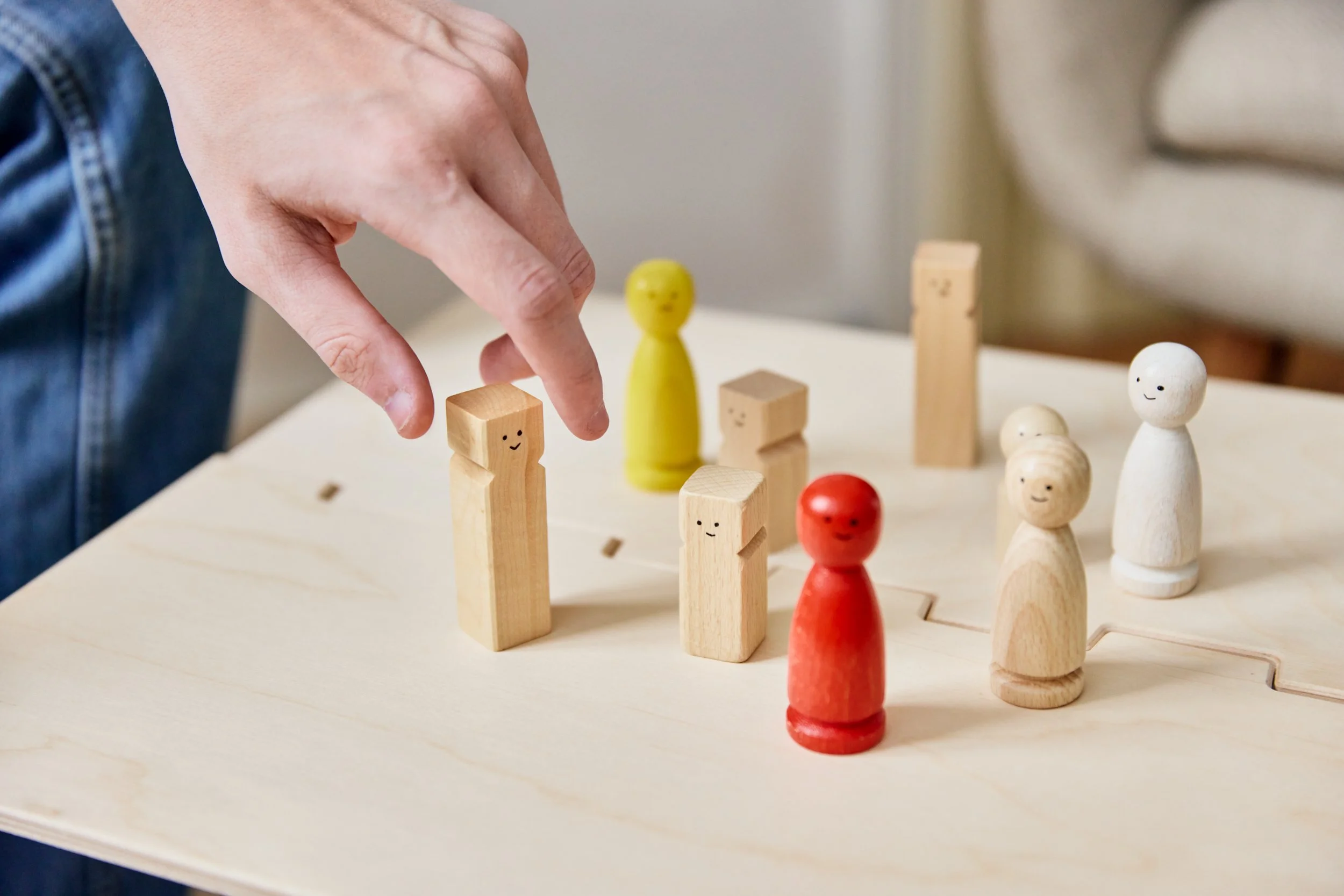 Hand bewegt einen kleinen Holzfiguren in einem Spiel, mit weiteren bunten Holzfiguren auf einem Tisch, Figuren mit lachenden Gesichtern, vermutlich Teil eines Gesellschaftsspiels.