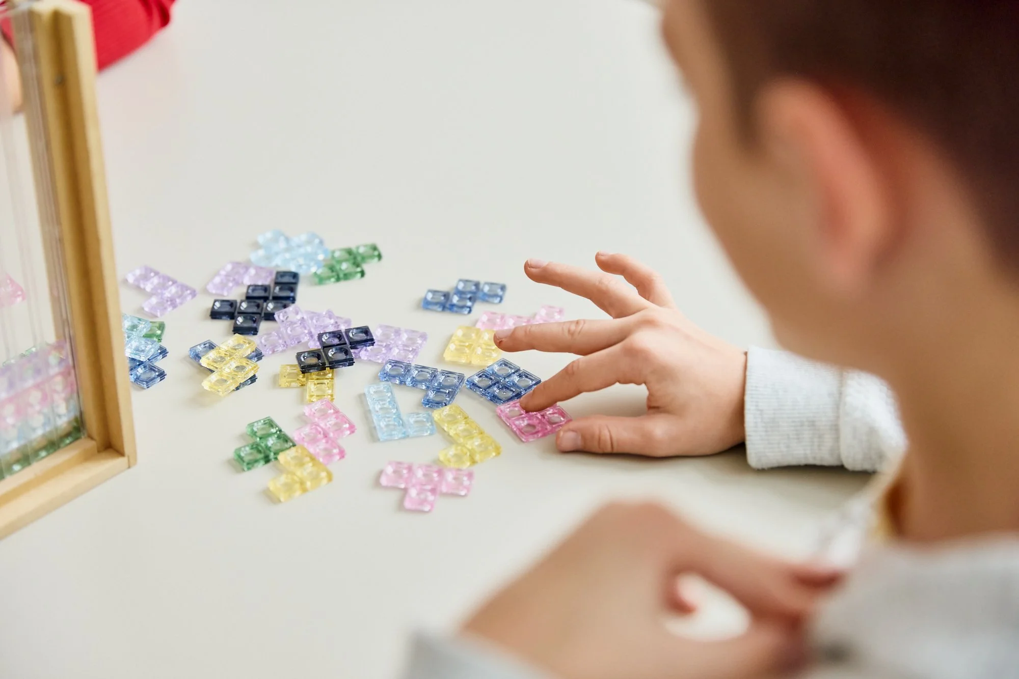 Ein Kind sortiert bunte, kleine, durchsichtig Katzenklappen auf einem Tisch, mit einem Holzrahmen voller weiterer ähnlicher Gegenstände auf der linken Seite.