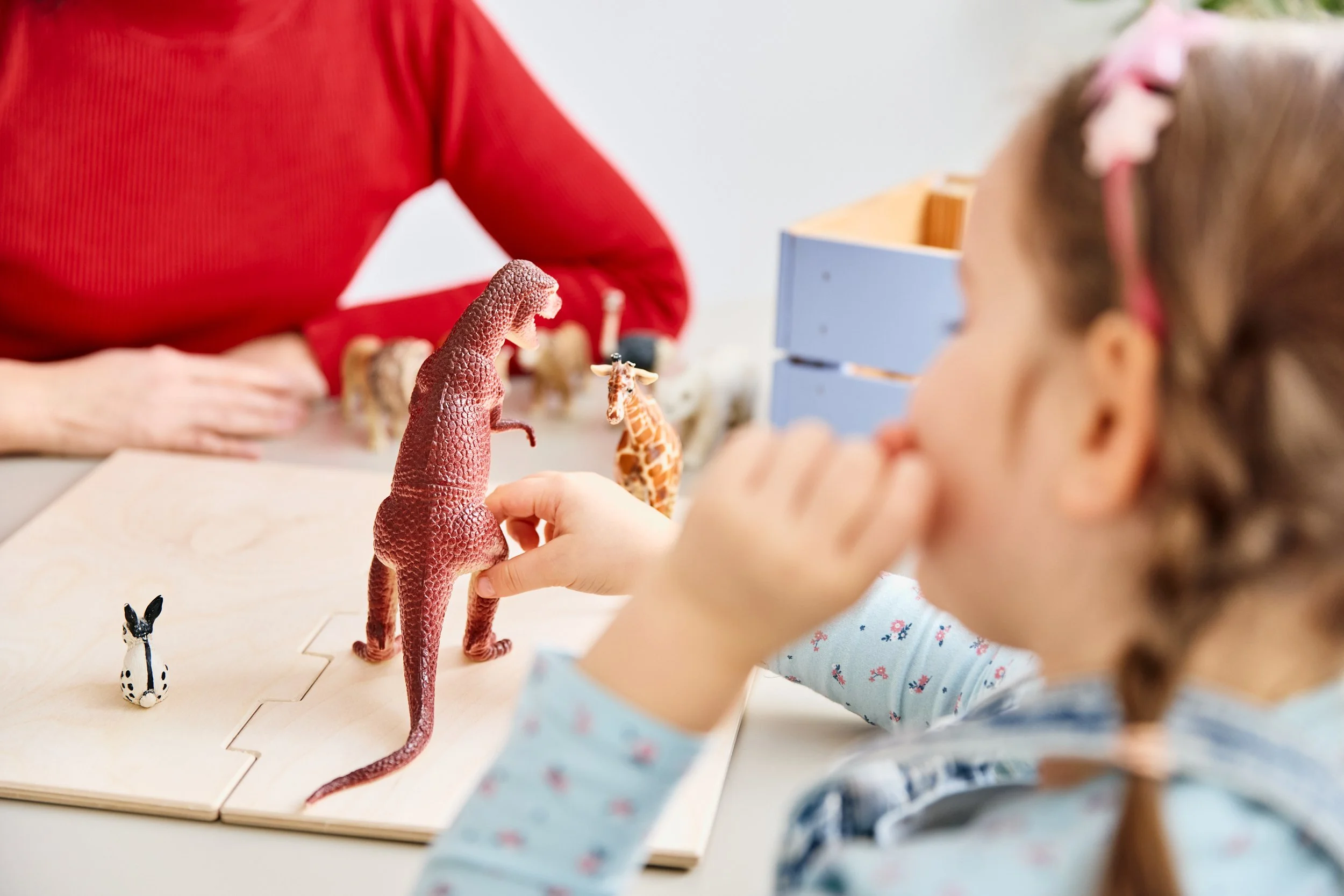 Ein Mädchen betrachtet Spielzeugtiere, darunter eine aufgebaute Dinosaurierfigur, während ein Erwachsener im Hintergrund sitzt.