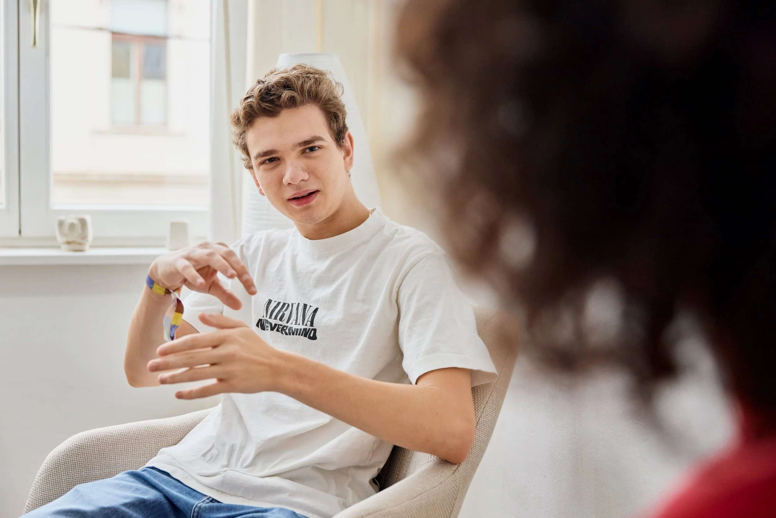 Ein junger Mann mit braunen, lockigen Haaren spricht in einem Gespräch. Er trägt ein weißes T-Shirt mit einem Nirvana-Logo und sitzt in einem hellen Raum vor einem Fenster, in dem Tageslicht einfällt.