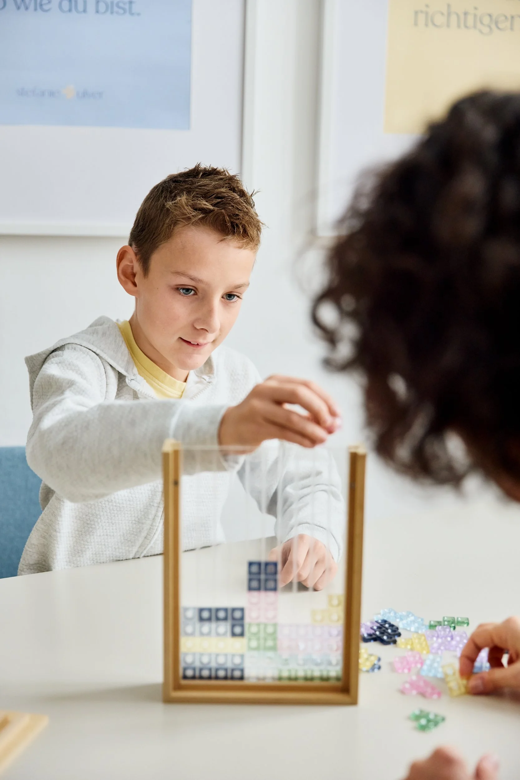 Ein Junge spielt ein Tangram-Puzzle mit bunten Steinen an einem Tisch in einem Klassenzimmer.