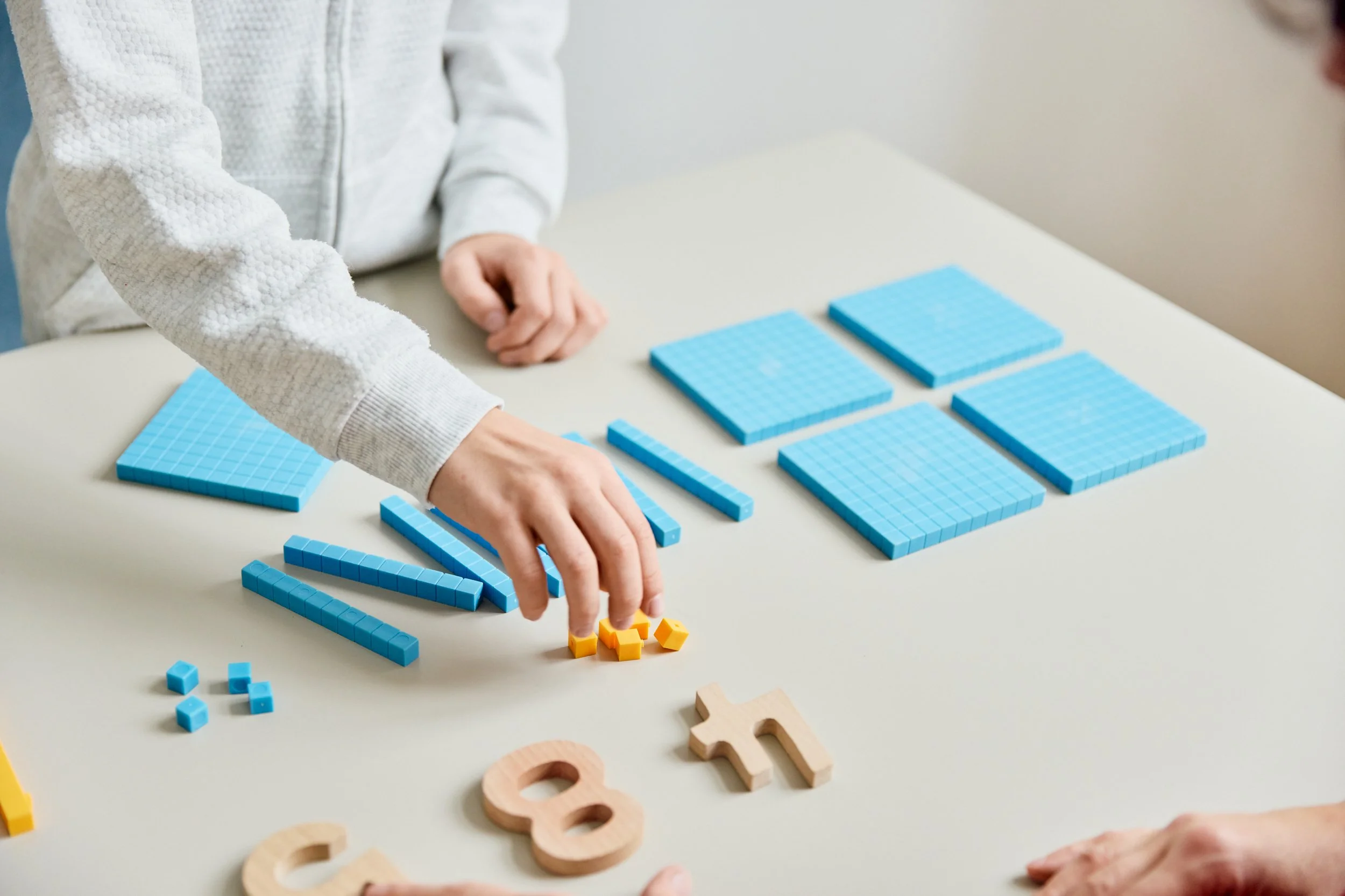 Nahaufnahme von Händen, die blaue Montessori-Lerntafeln, kleine gelbe und blaue Würfel sowie Holzbuchstaben auf einem weißen Tisch anordnen.