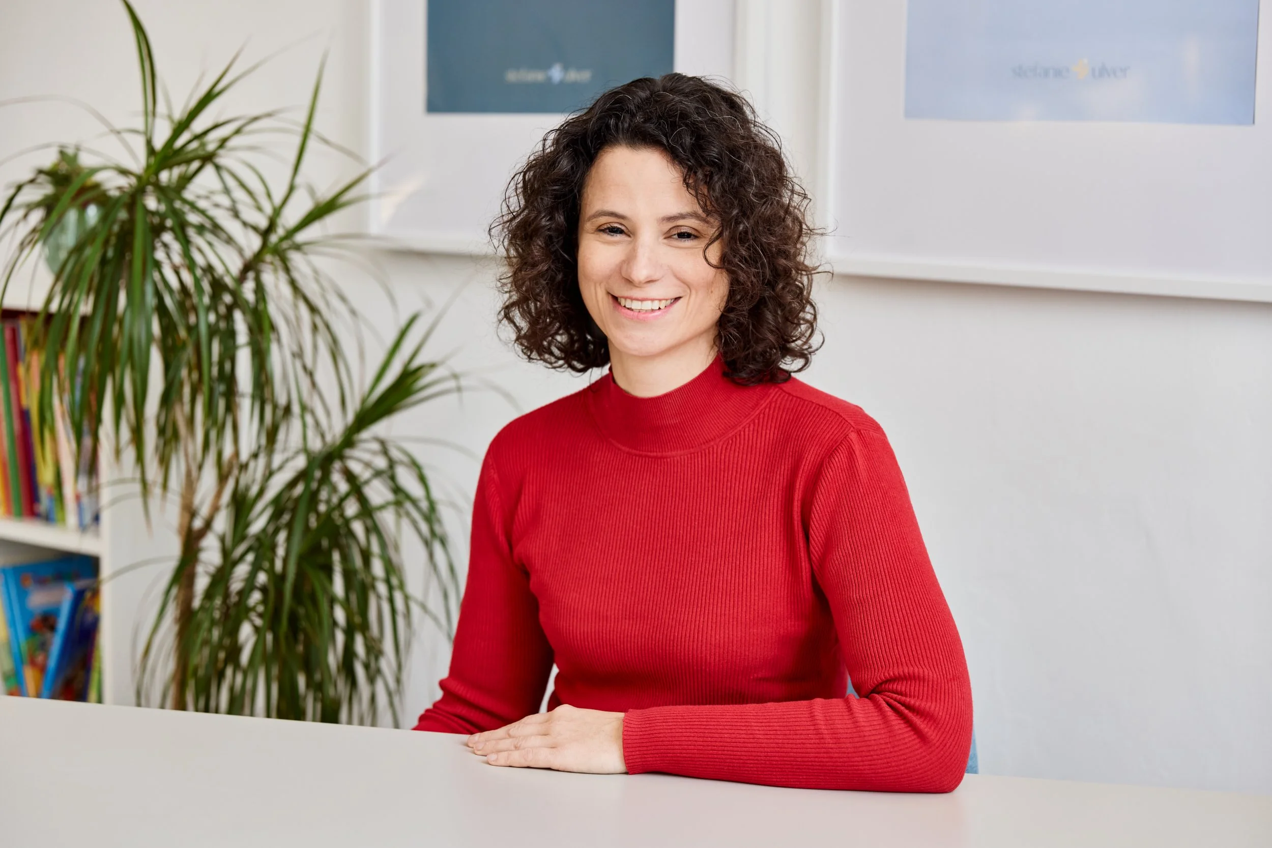 Eine lächelnde Frau mit lockigem braunem Haar sitzt an einem weißen Tisch vor einem Bücherregal und einer Pflanze, im Hintergrund ein Poster auf der Wand.
