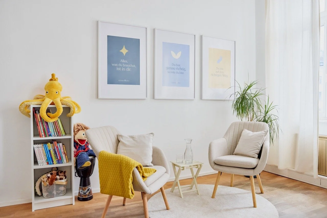 Gemütliches Wohnzimmer mit weißen Wänden, zwei Ohrensessel, eine kleine weiße Beistelltisch mit einer Glas Karaffe, eine grüne Zimmerpflanze, Bilder mit Zitaten an der Wand, ein Bücherregal mit Büchern und Spielzeug, großer Fenster mit weißen Vorhängen.