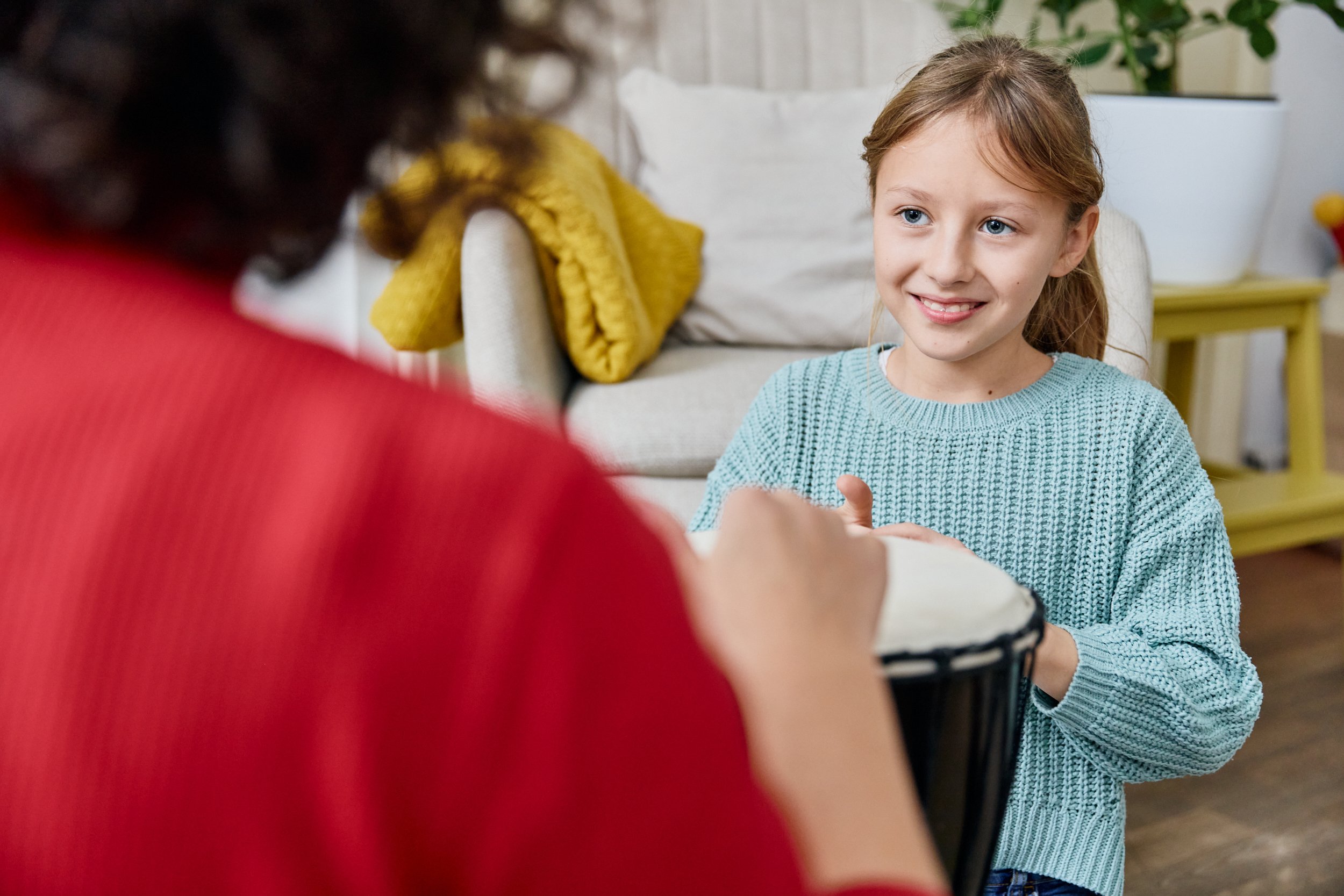 Ein Mädchen mit braunen Haaren spielt auf einem Tamburin, während eine Person mit rotem Pullover im Vordergrund spielt. Das Mädchen lächelt freundlich und befindet sich in einem Wohnzimmer.