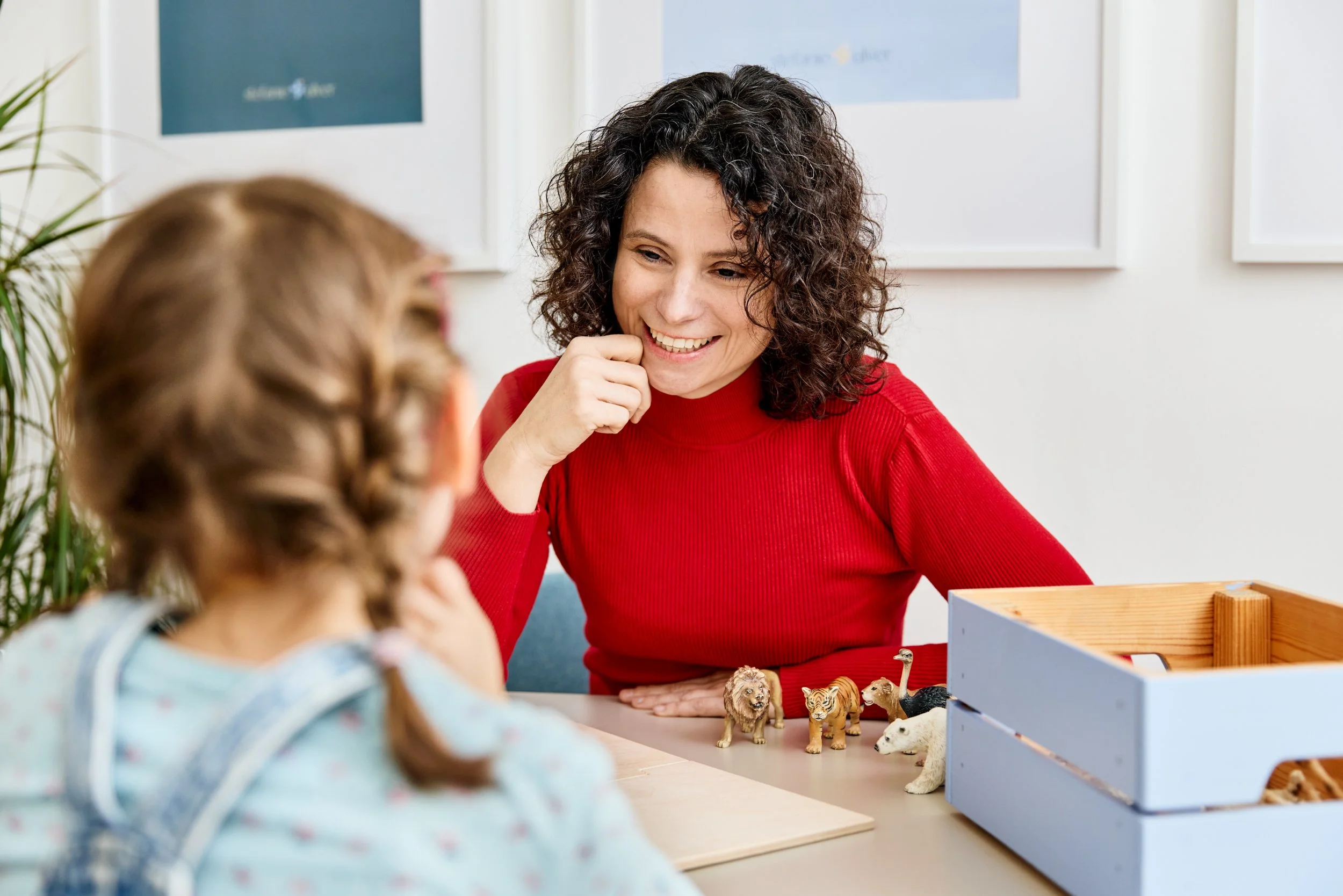 Eine Frau im roten Pullover spielt mit einem kleinen Mädchen und Tierfiguren auf einem Tisch.