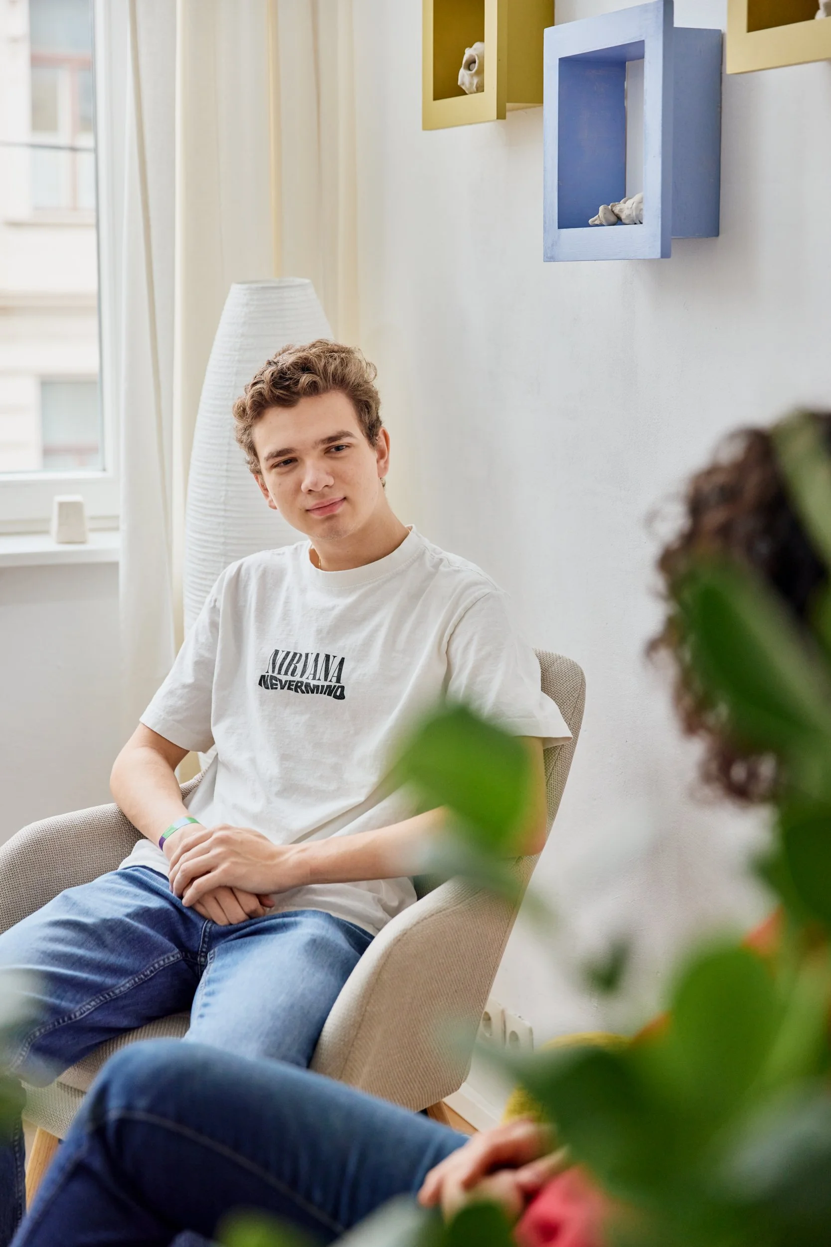 Ein junger Mann sitzt in einem beige Lounge-Stuhl in einem hellen Raum, trägt ein weißes Nirvana-T-Shirt und blickt in die Kamera. Im Vordergrund sind verschwommene grüne Blätter einer Pflanze sichtbar.