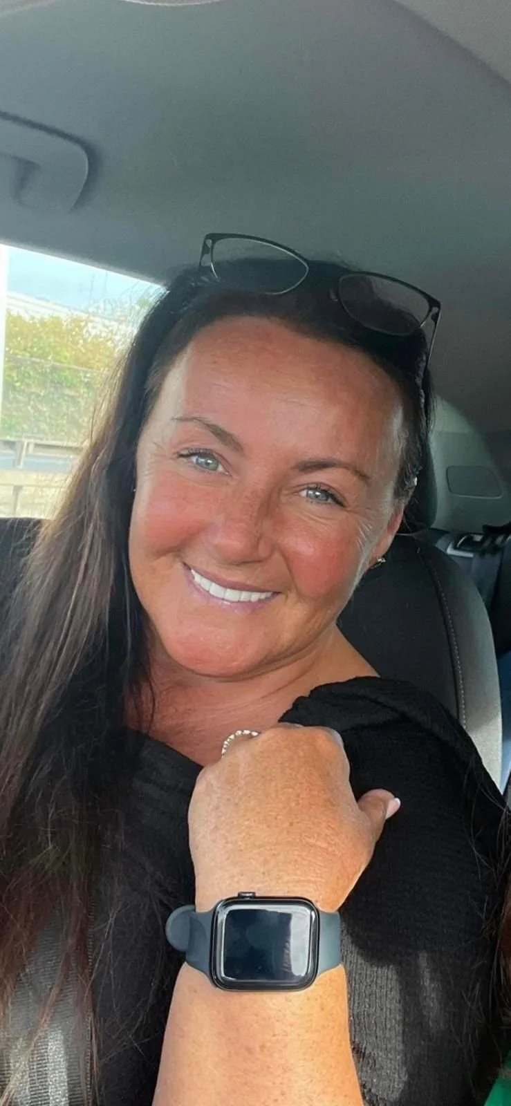 A woman taking a selfie outdoors near the water, wearing large sunglasses and a white bikini, with a rocky shoreline and boat in the background.