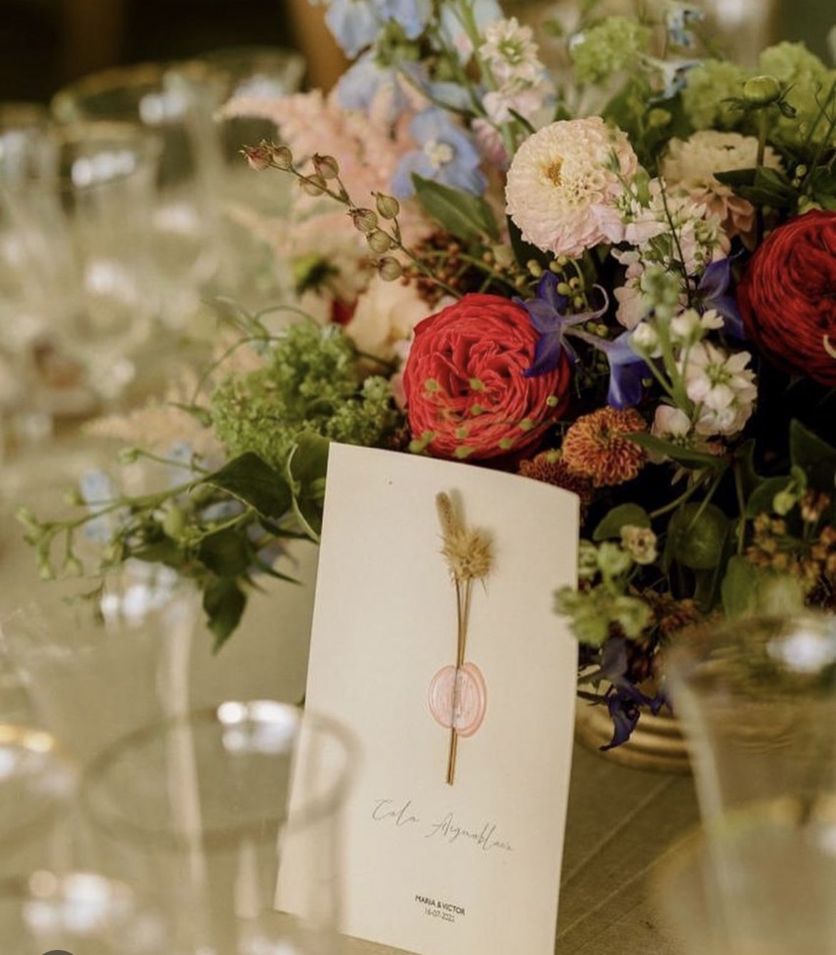 Ramos de flores en tonos rosados, rojos, blancos y azules en un centro de mesa, con una tarjeta en primer plano y copas de vino en el fondo.