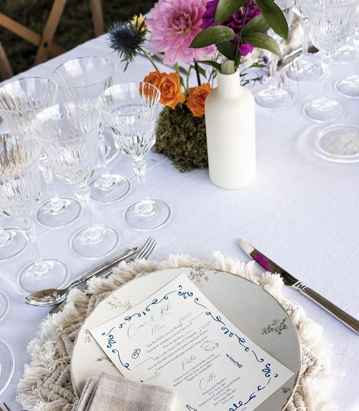 Mesa decorada con varios copas de cristal, un florero blanco con flores de colores, una invitación y utensilios para comer sobre un mantel blanco.