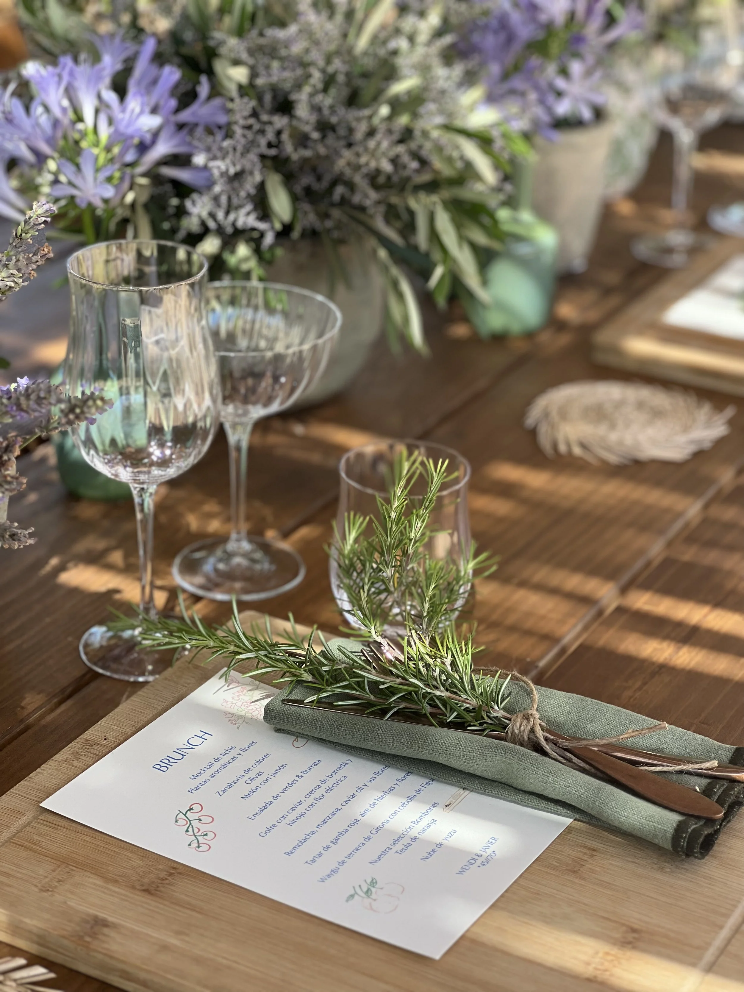 Mesa de madera decorada con flores moradas, vasos de vidrio y un ramo de romero en un rollo de tela. Hay un menú en la mesa y plumas de pluma de ave. Iluminación natural con sombras suaves.