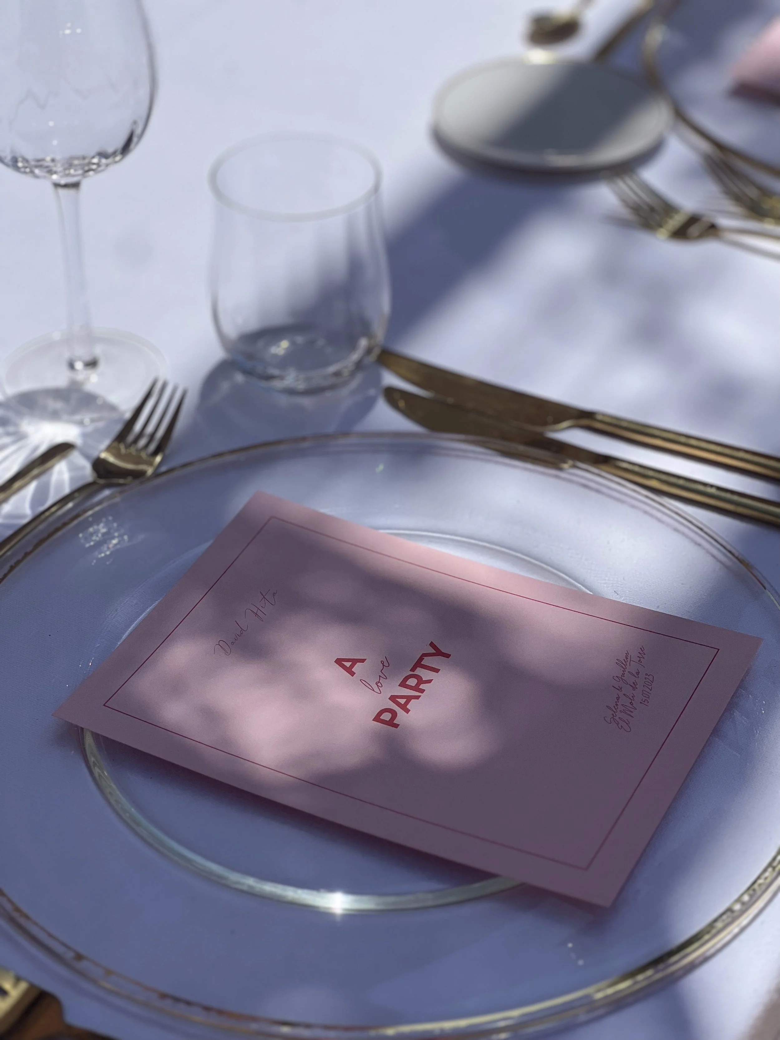 Mesa elegant con plato de cristal, collage, la invitación que dice 'A little party' y cubiertos dorados, en la mesa hay un vaso de agua, un vaso de vino y un platito pequeño.