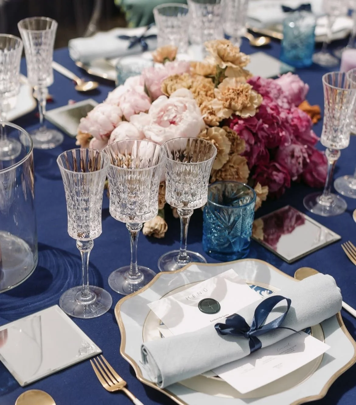 Mesa decorada con flores y cristalería, tonos de azul, rosa y beige, con menús y cubertería elegante para evento formal.