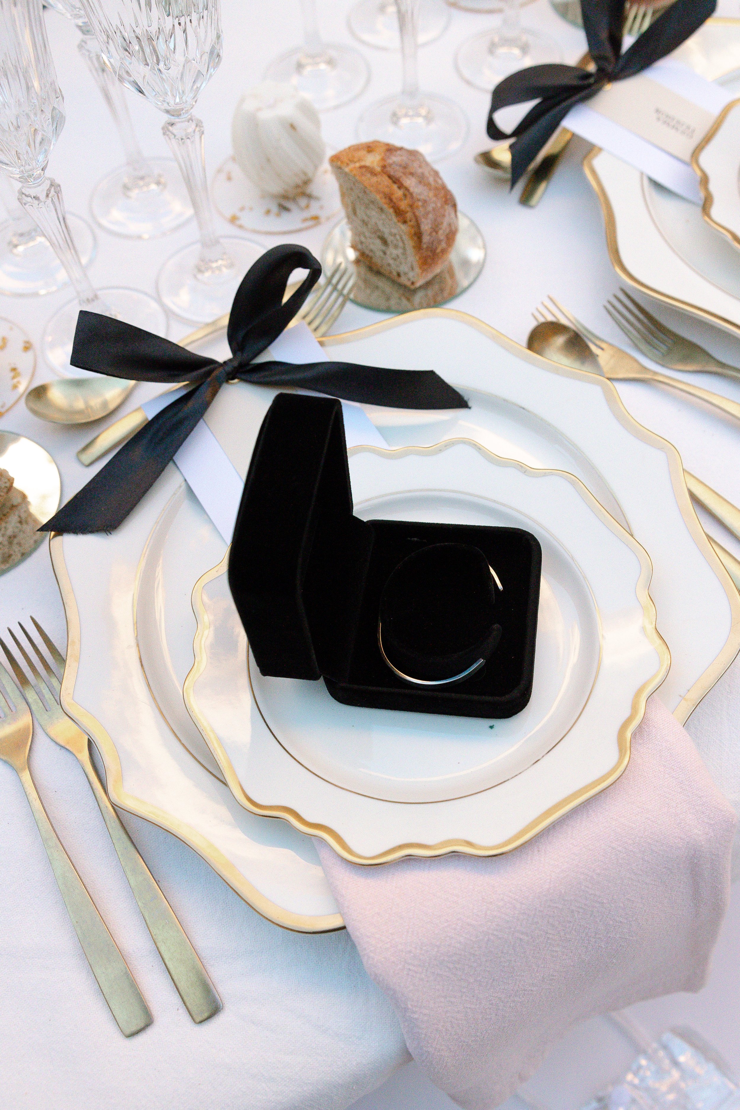 Un plato de platos elegantes con un anillo de compromiso en una caja negra en el centro, en una mesa decorada con copas de cristal, cubiertos dorados, un pan y una etiqueta con la palabra 'COMPRAR' y un lazo negro.
