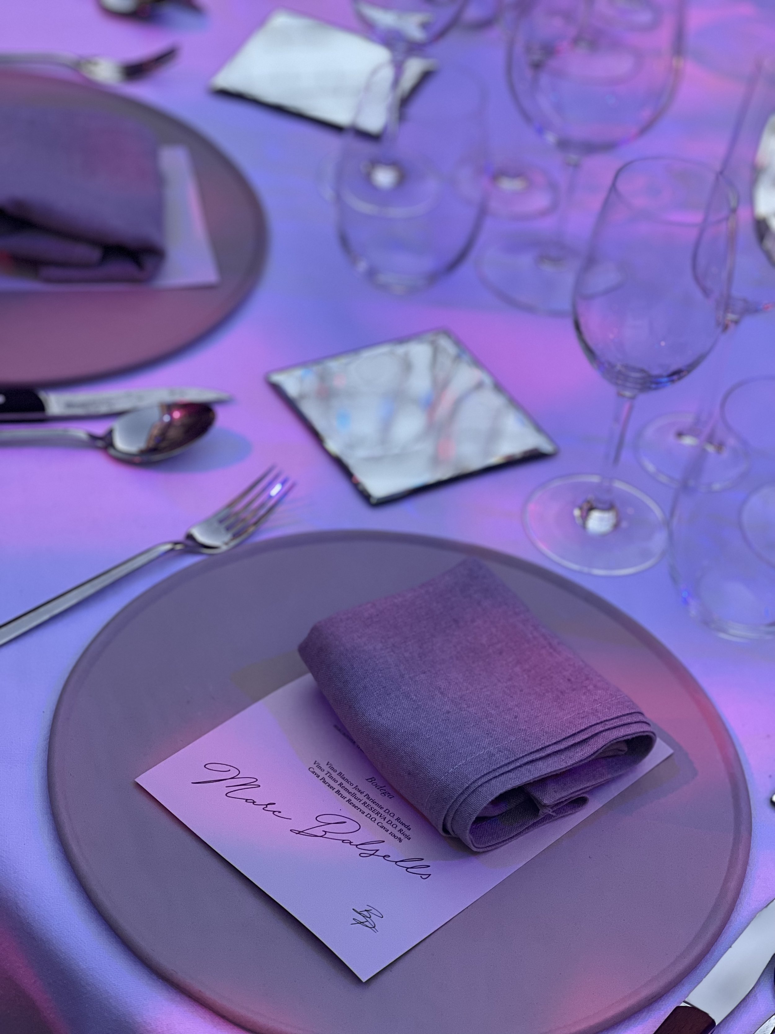 Mesa decorada para un evento con platos, cubiertos, copas y un servilleta gris en un lugar con iluminación azul y rosa